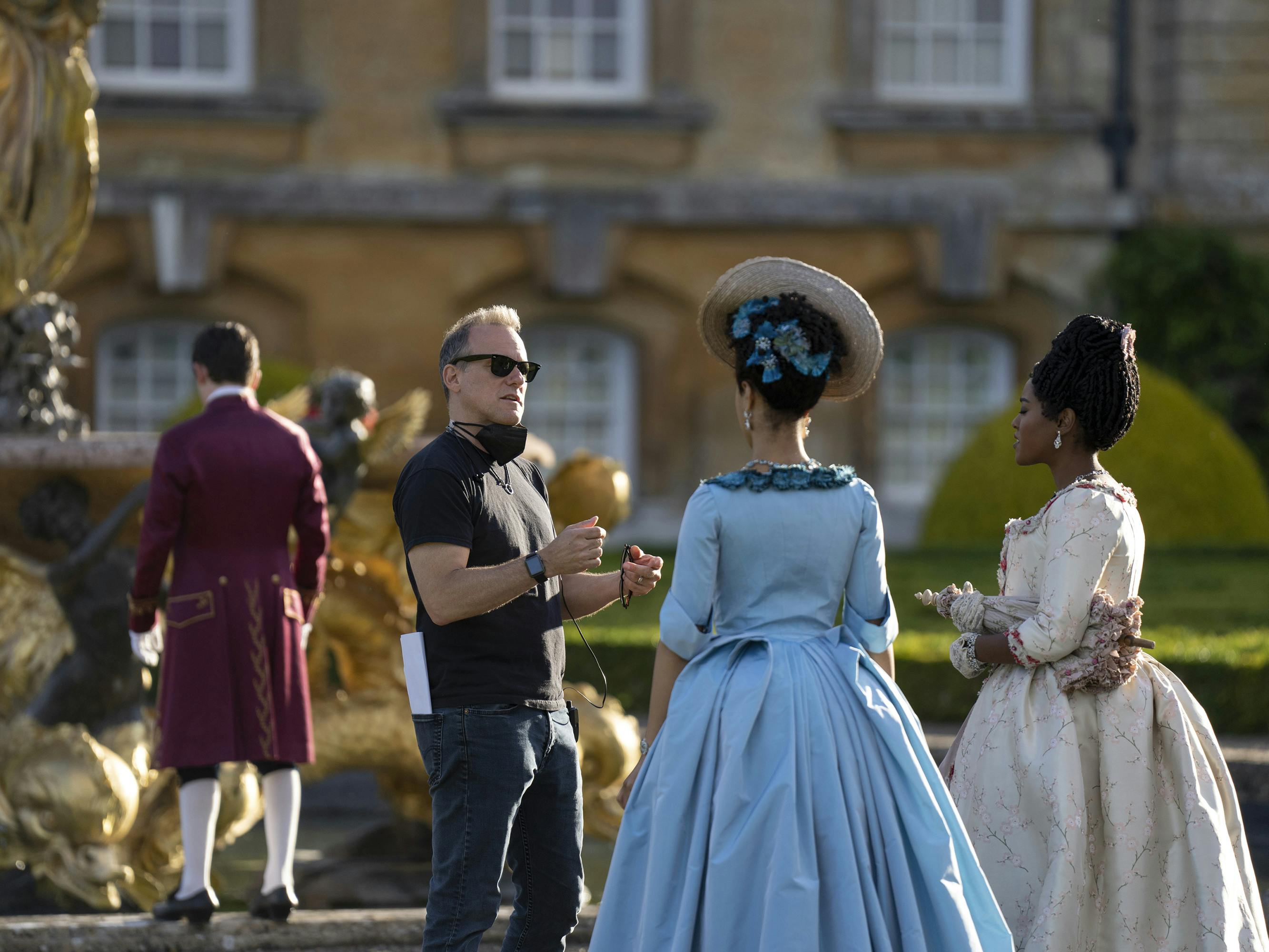 Tom Verica, India Amarteifio, and Arsema Thomas stand around on set. Amarteifio wears a blue dress and headpiece, Arsema wears white, and Verica wears black with matching shades.
