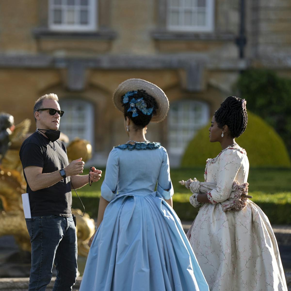 Tom Verica, India Amarteifio, and Arsema Thomas stand around on set. Amarteifio wears a blue dress and headpiece, Arsema wears white, and Verica wears black with matching shades.