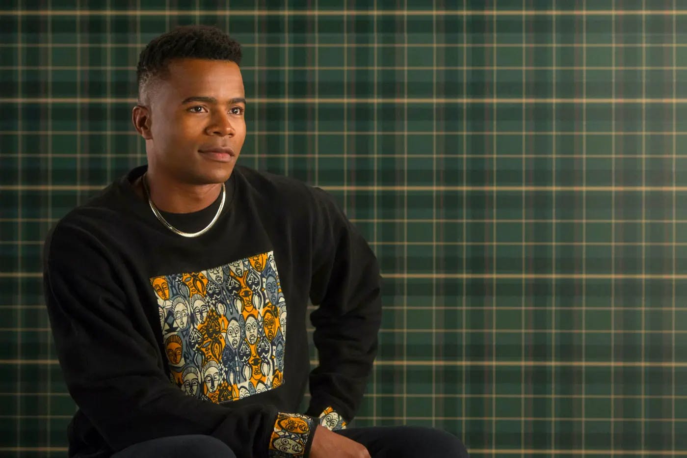 Reggie Green (Marque Richardson) sits relaxed in front of a green checked background.  He wears a silver necklace, dark blue jeans, and a black sweatshirt with sections on the front and cuffs that feature a grey and yellow abstract print of faces.