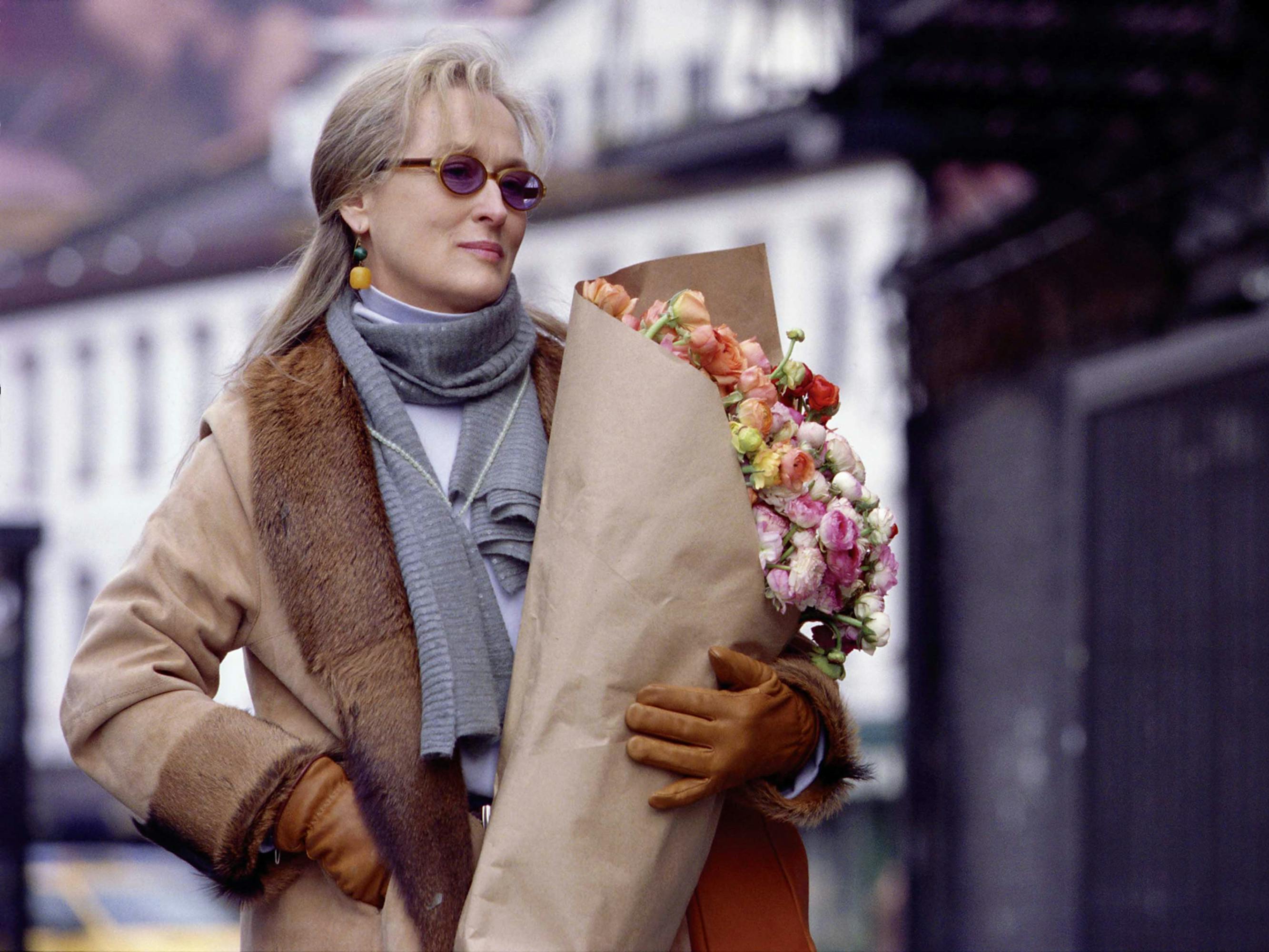 Meryl Streep carries a large bouquet of flowers wrapped in brown paper. She wears sunglasses, a blue scarf, and a tan jacket with a fur collar. 