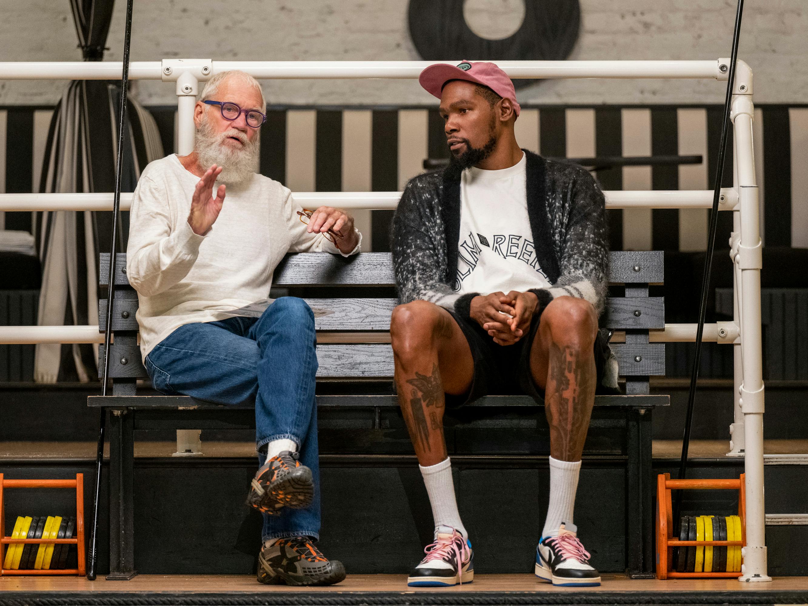 David Letterman and Kevin Durant sit on a bench. Letterman wears jeans and an oatmeal sweater. Durant wears sneakers and a baseball hat.