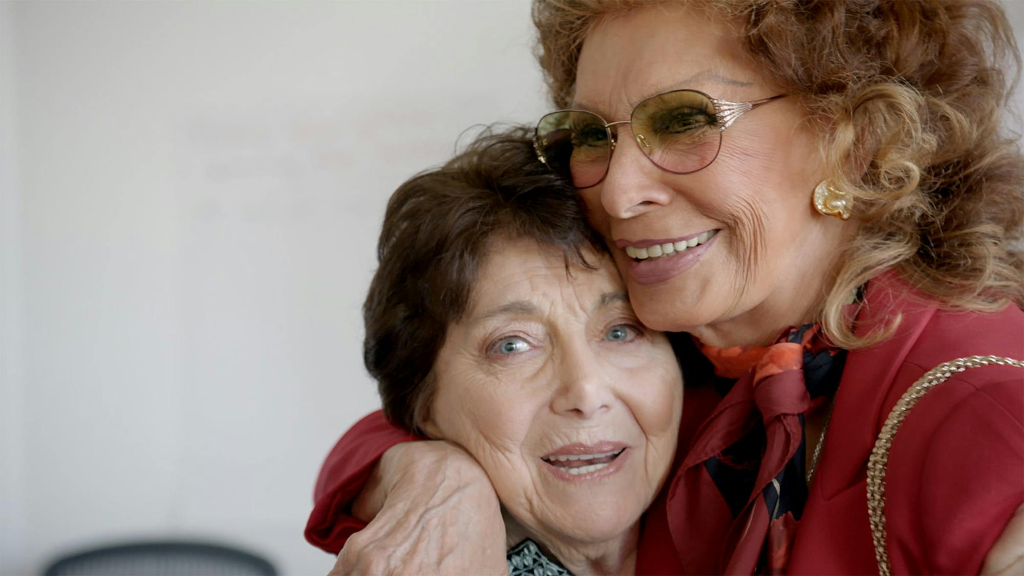 A close-up of Kulik and Loren as they embrace. Kulik looks straight to camera. Loren wears a red blazer, scarf, and sunglasses.