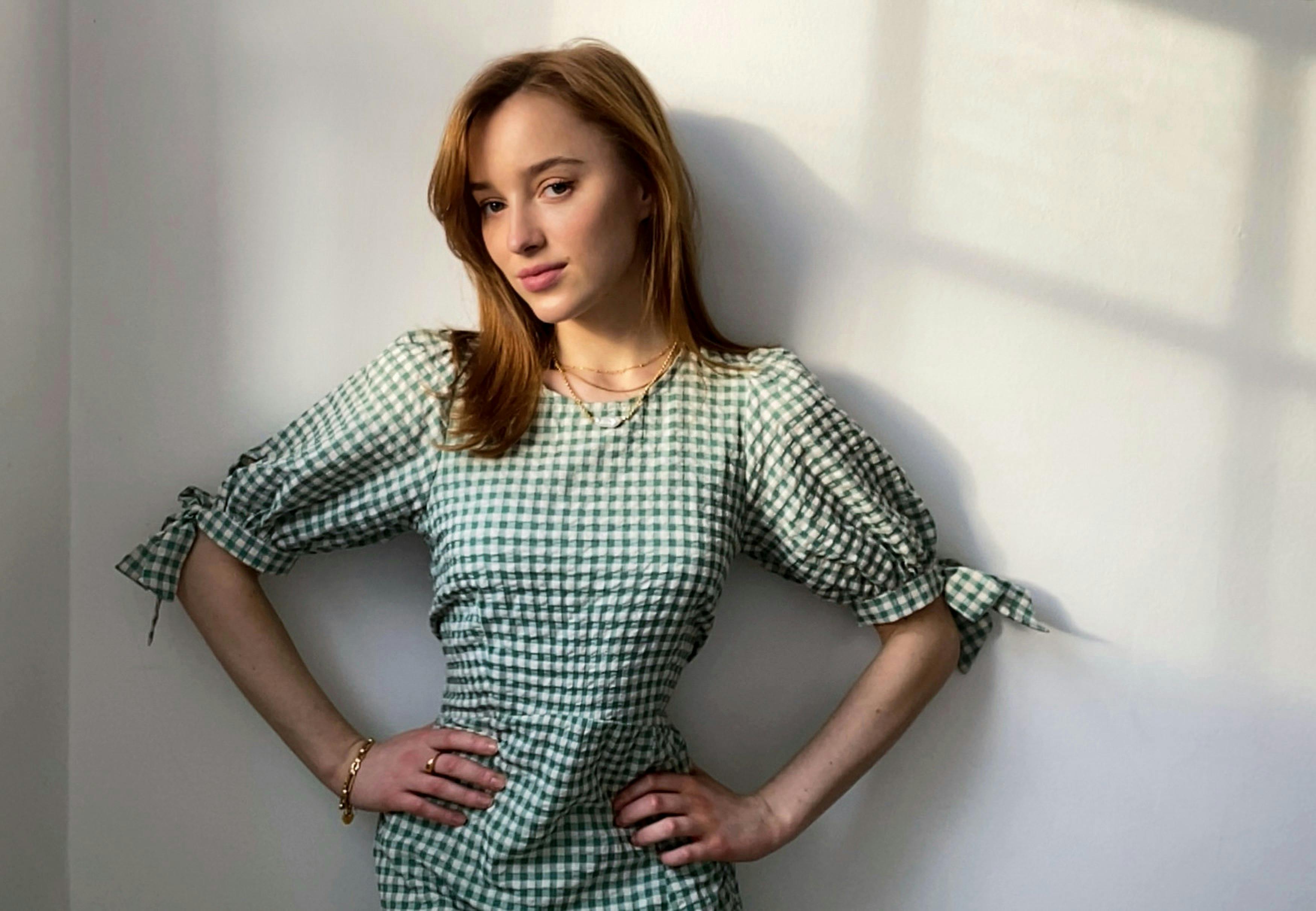 The actress stands with her hands on her waist in a green gingham dress. Her face is illuminated by some golden hour light. 