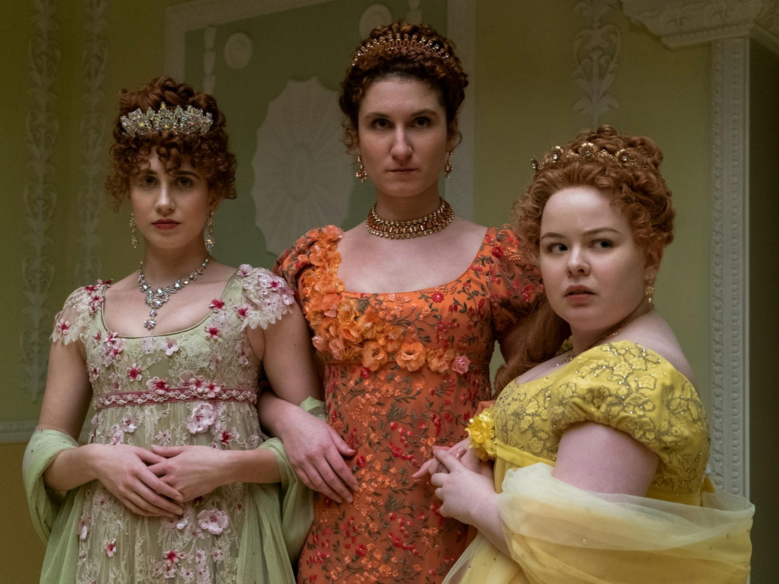 Penelope Featherington (Nicola Coughlan) stands beside her sisters. Coughlan wears a yellow gown, while one sister wears orange and the other green with pink flowers. They look judgemental and intimidating with their arms linked together.