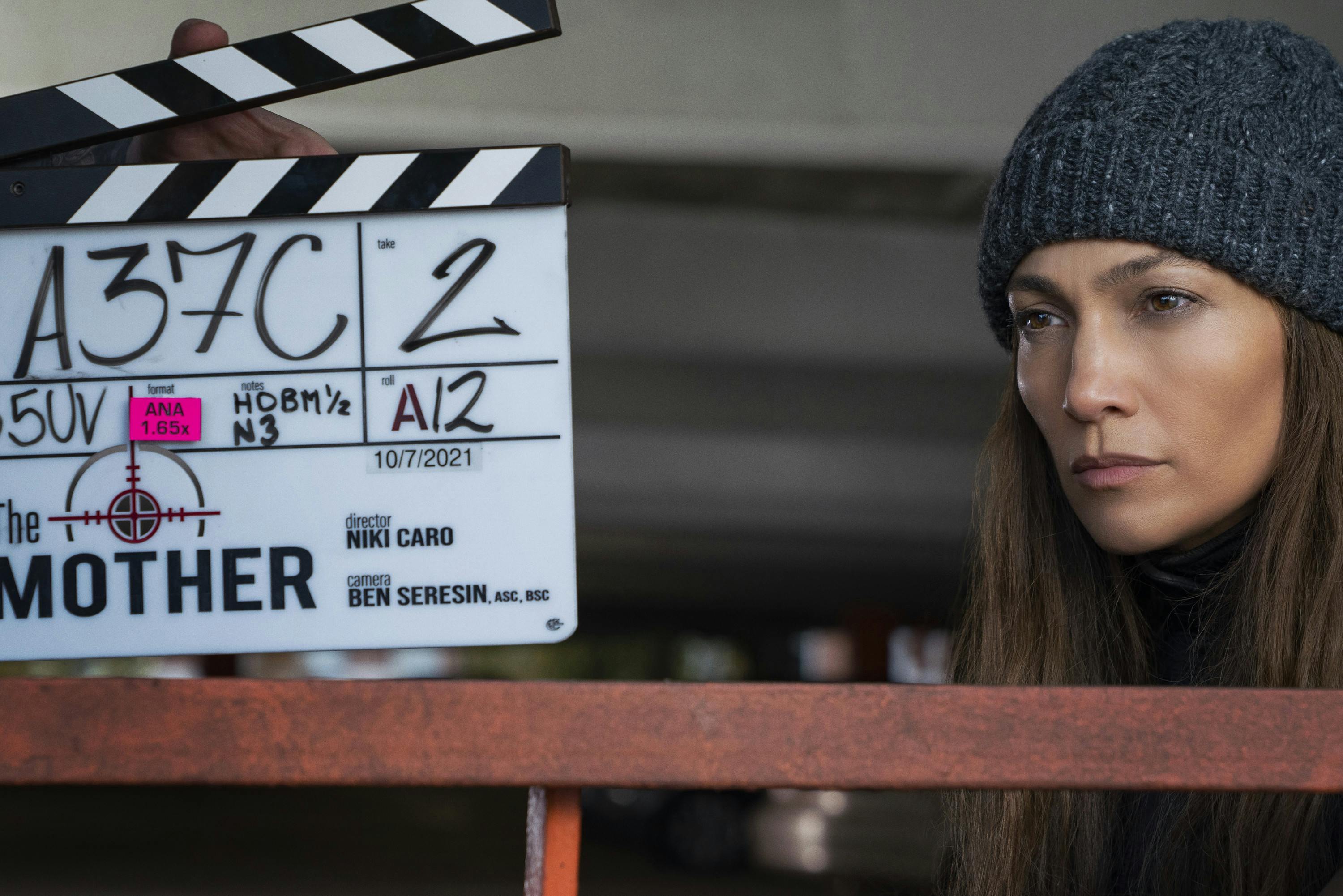 Jennifer Lopez wears a grey beanie and stands behind the scenes of The Mother. In front of her face is a clapperboard. 