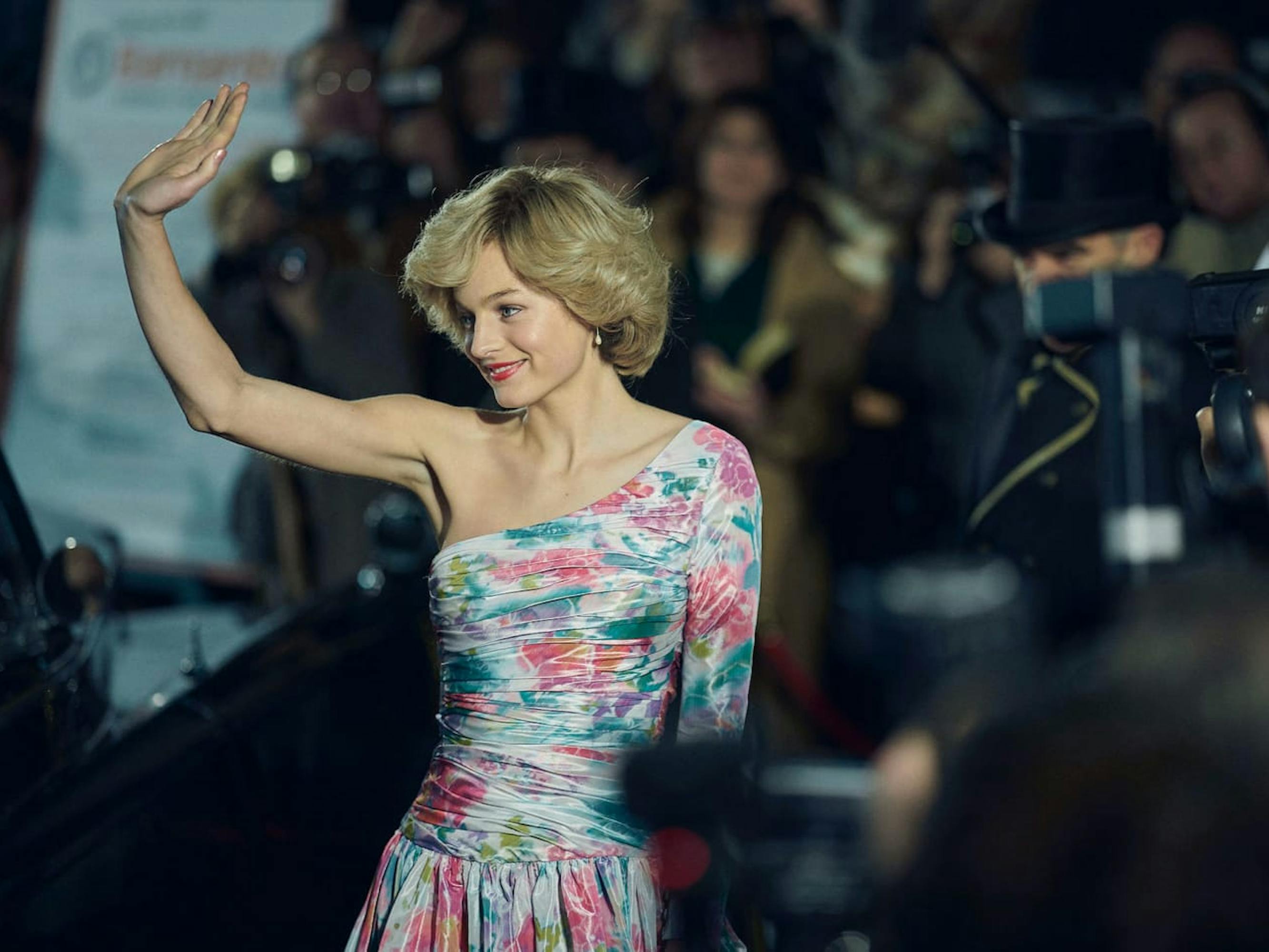 Diana (Emma Corrin) waves to the crowd, surrounded by people and cameras. She wears an off the shoulder floral dress.
