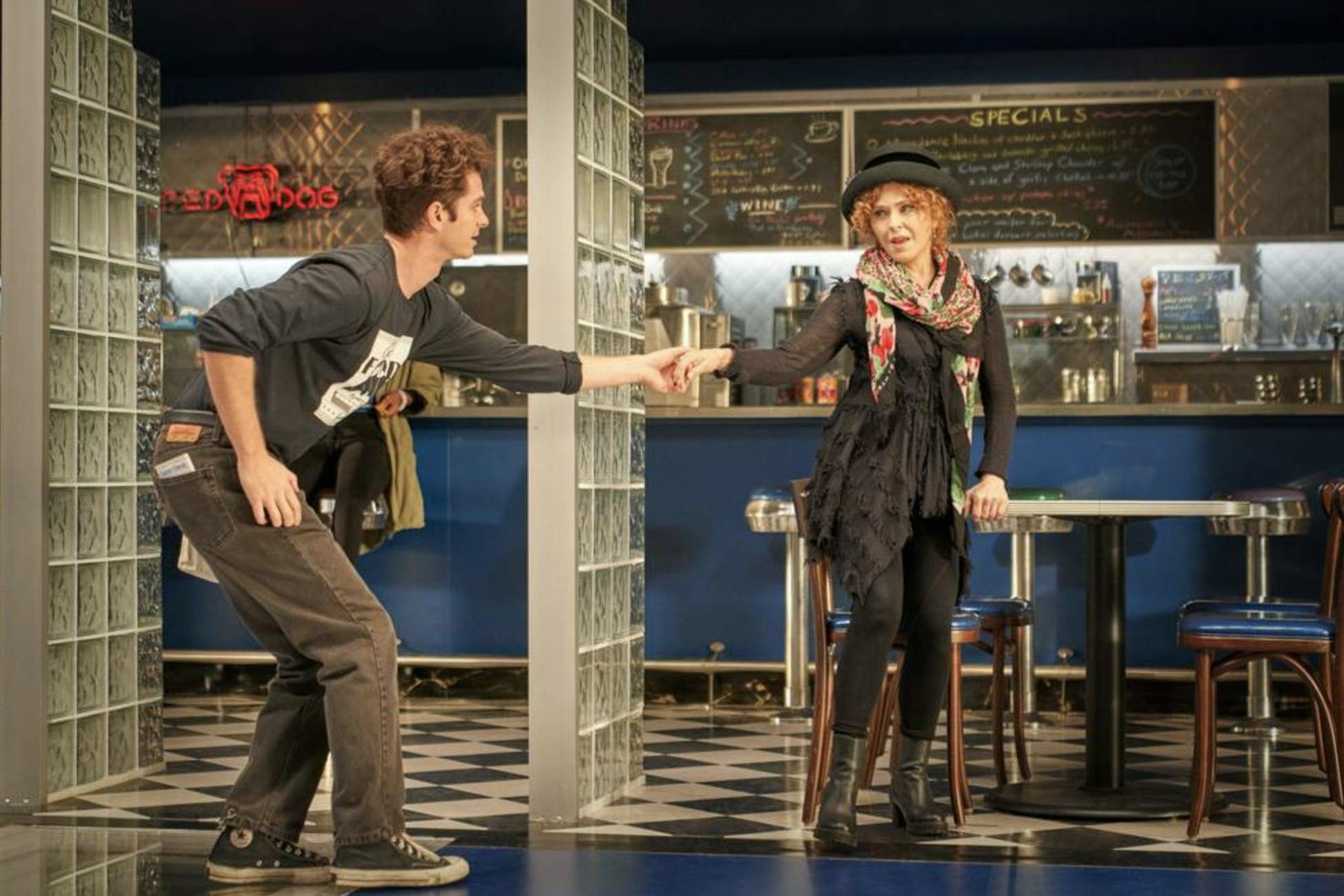 Andrew Garfield reaches for Bernadette Peters's hand in the Moondance Diner. Dishes are written in colorful chalk on the blackboard behind the counter. Garfield wears brown pants, converse, and a black Moondance Diner shirt. Peters wears a black ensemble with a colorful scarf, and a dark hat. 