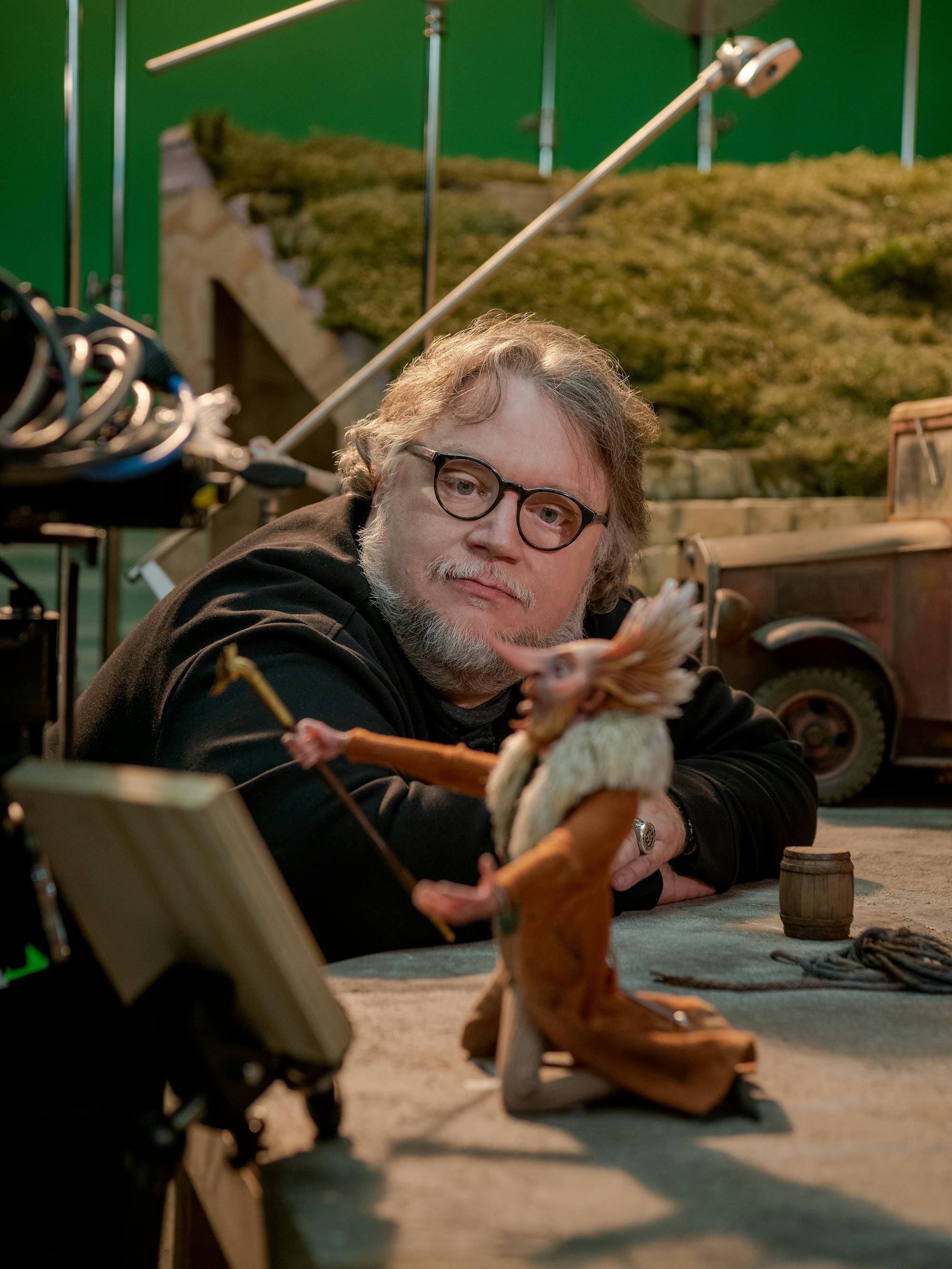 Guillermo del Toro examines the Count Volpe (Christoph Waltz) puppet with a pensive expression. Behind them is a grassy set.