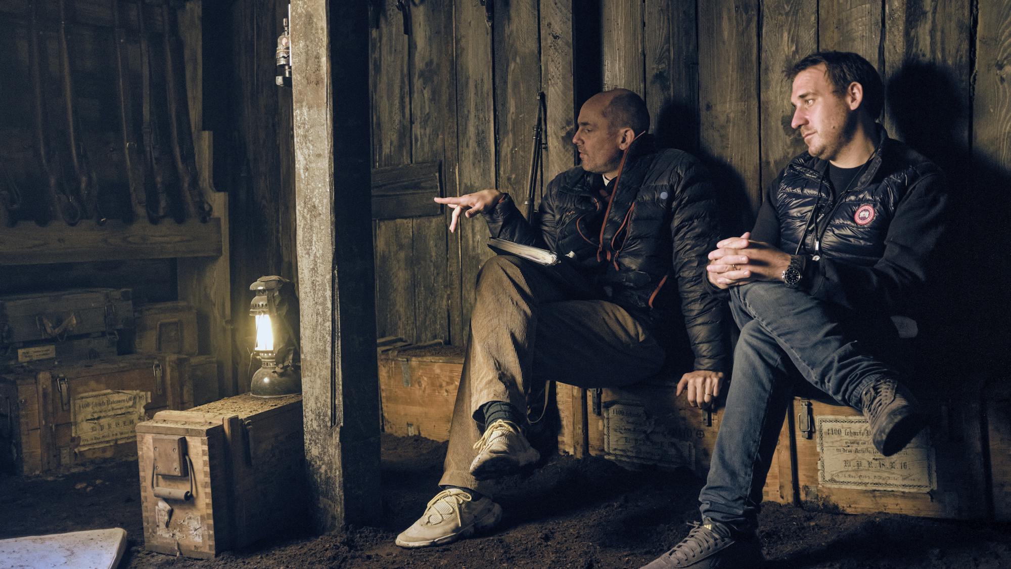 Edward Berger and James Friend sit together in a dirty wood room lit by a latern.