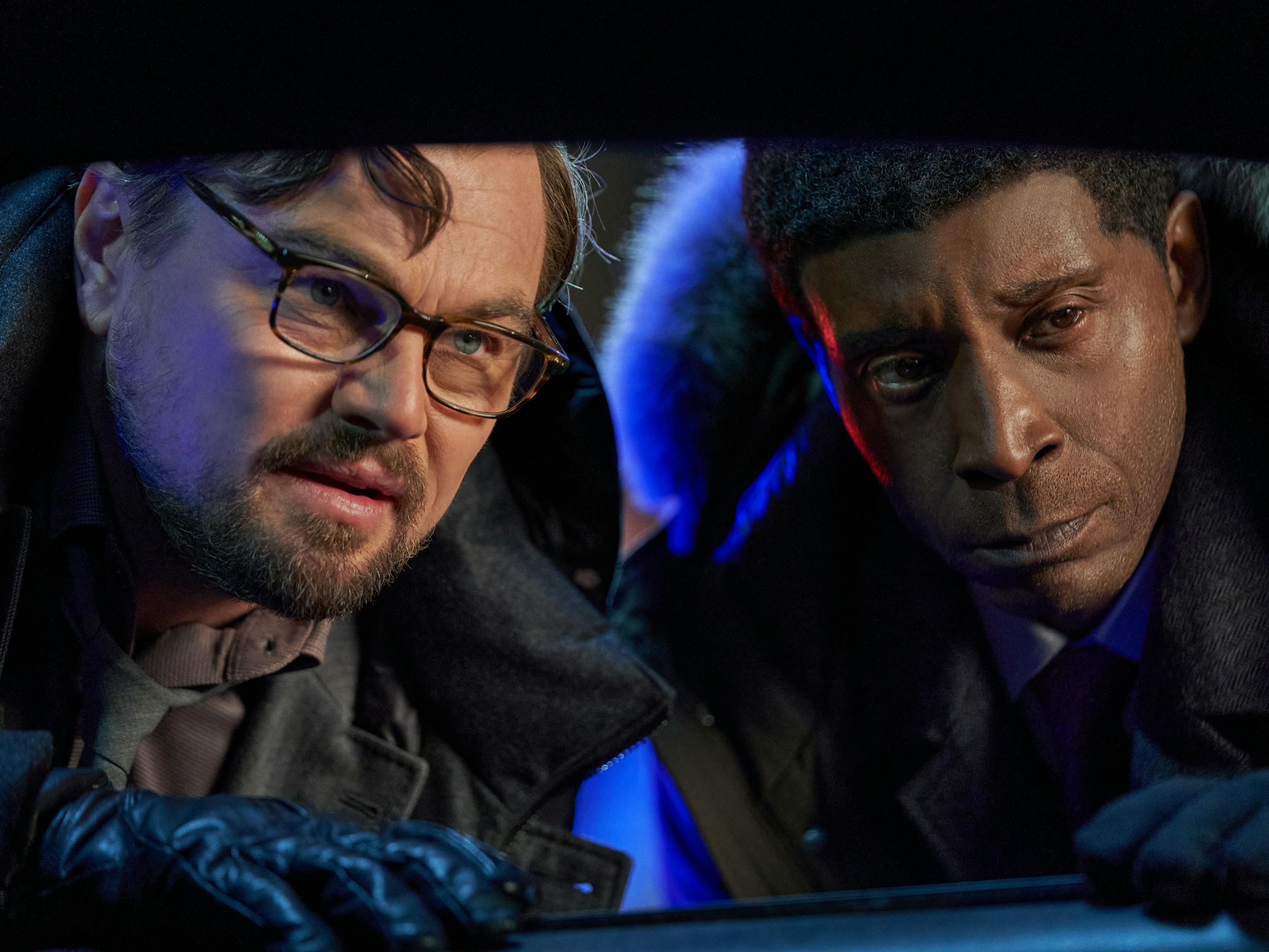 Leonardo DiCaprio and Rob Morgan look through a window. DiCaprio wears a dark jacket and dark gloves. Morgan wears a dark jacket with a furry hood.