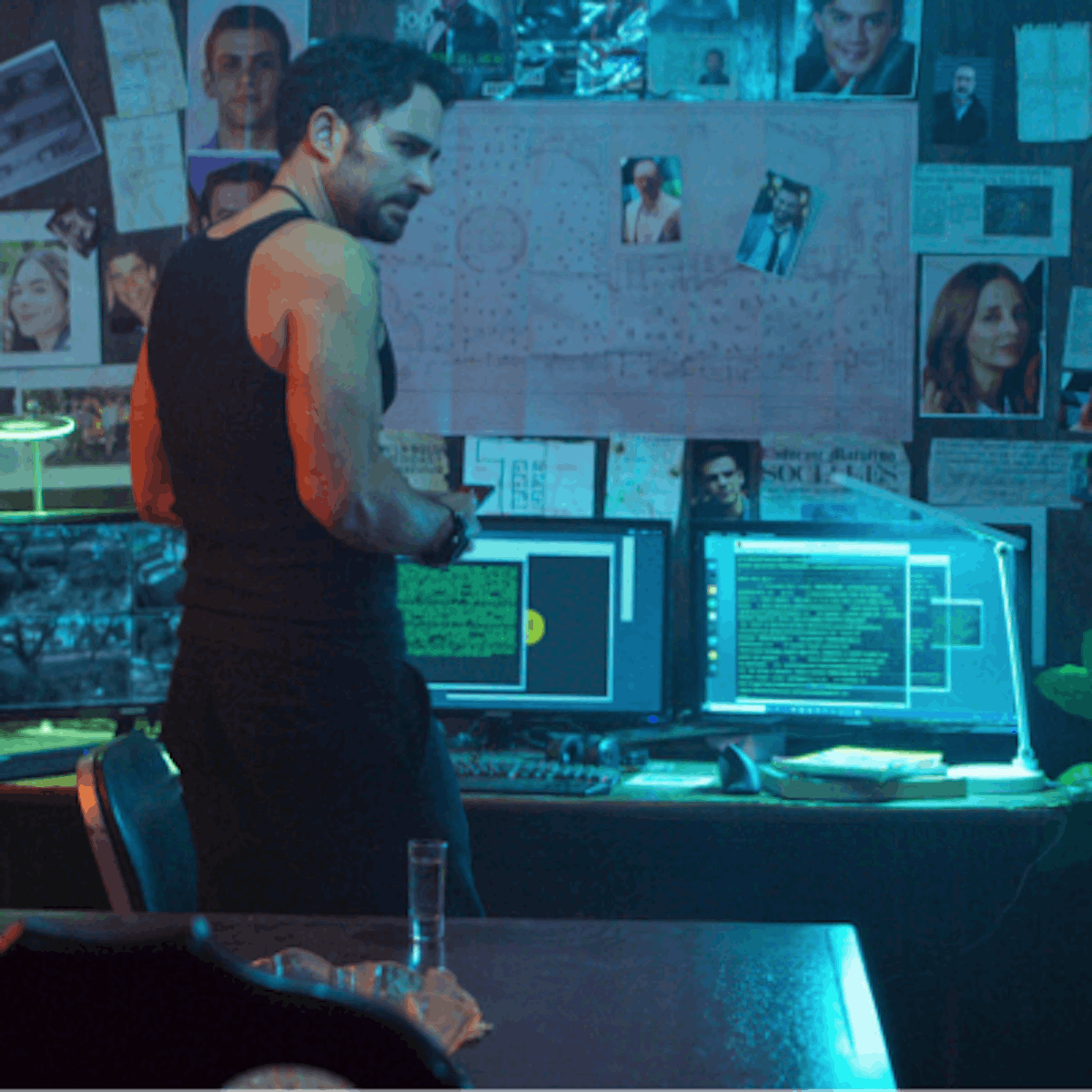 A man stands in front of a display of computers, pictures taped to the wall, and a map. He wears a black tank top and black pants, and has a sinister look on his face. There are plants flanking the table, and the room is lit with eerie blue light.