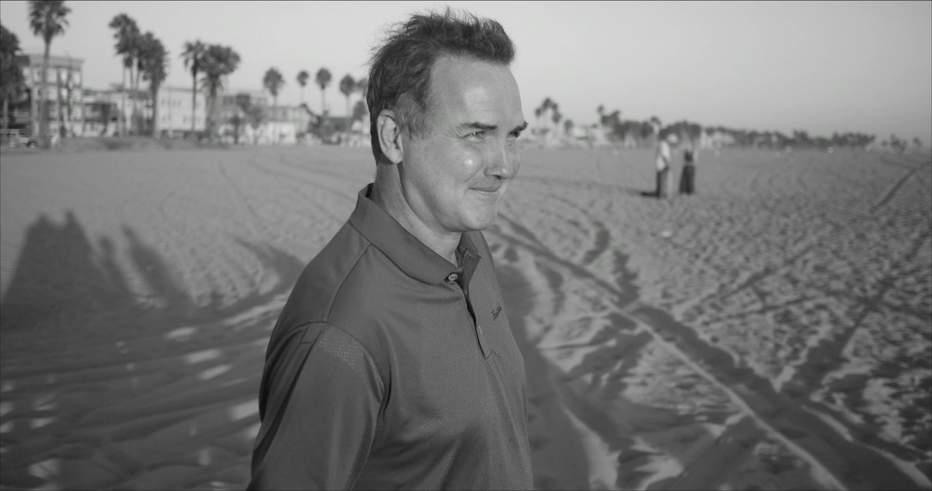 Norm Macdonald stands on a beach, his face lit by the sun.