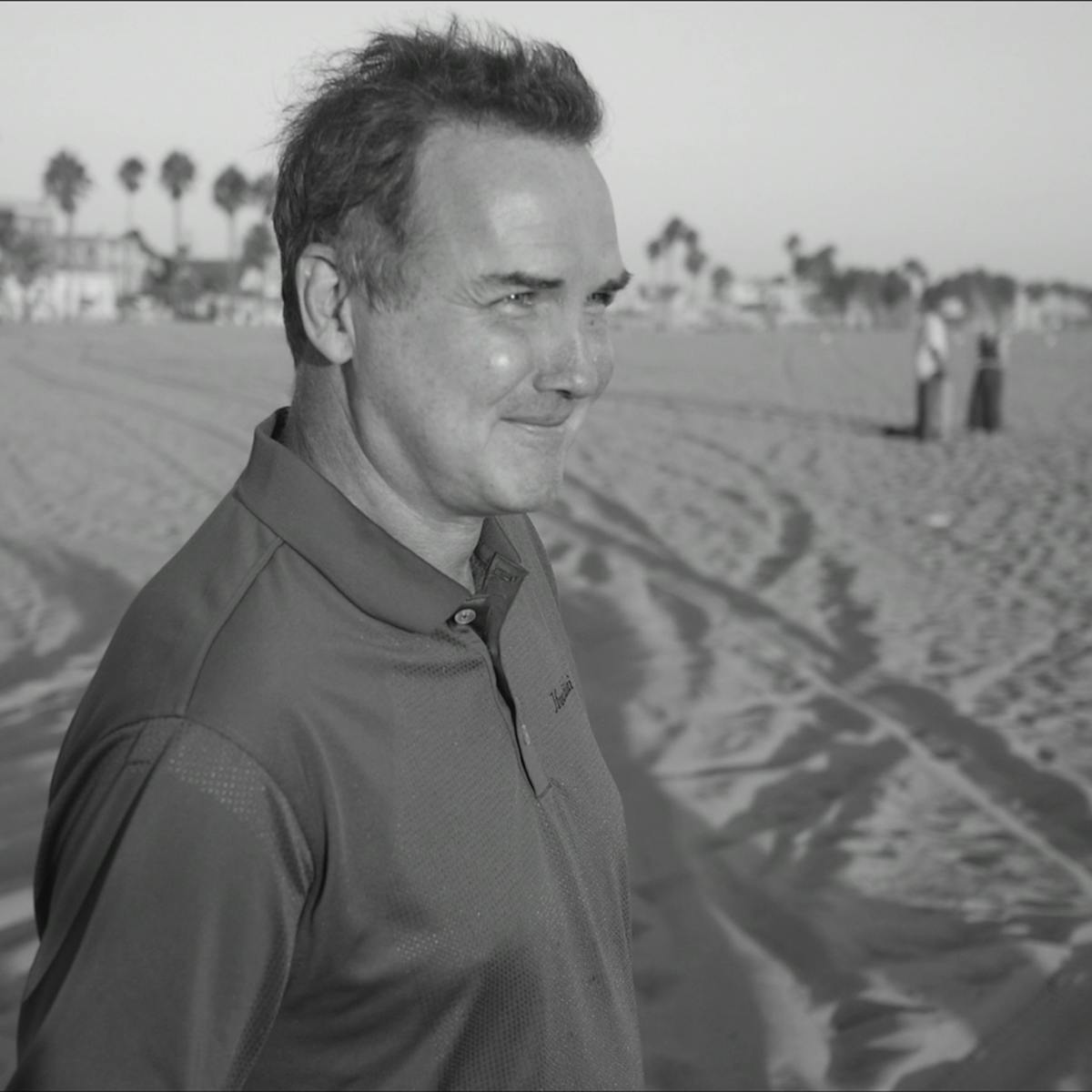 Norm Macdonald stands on a beach, his face lit by the sun.