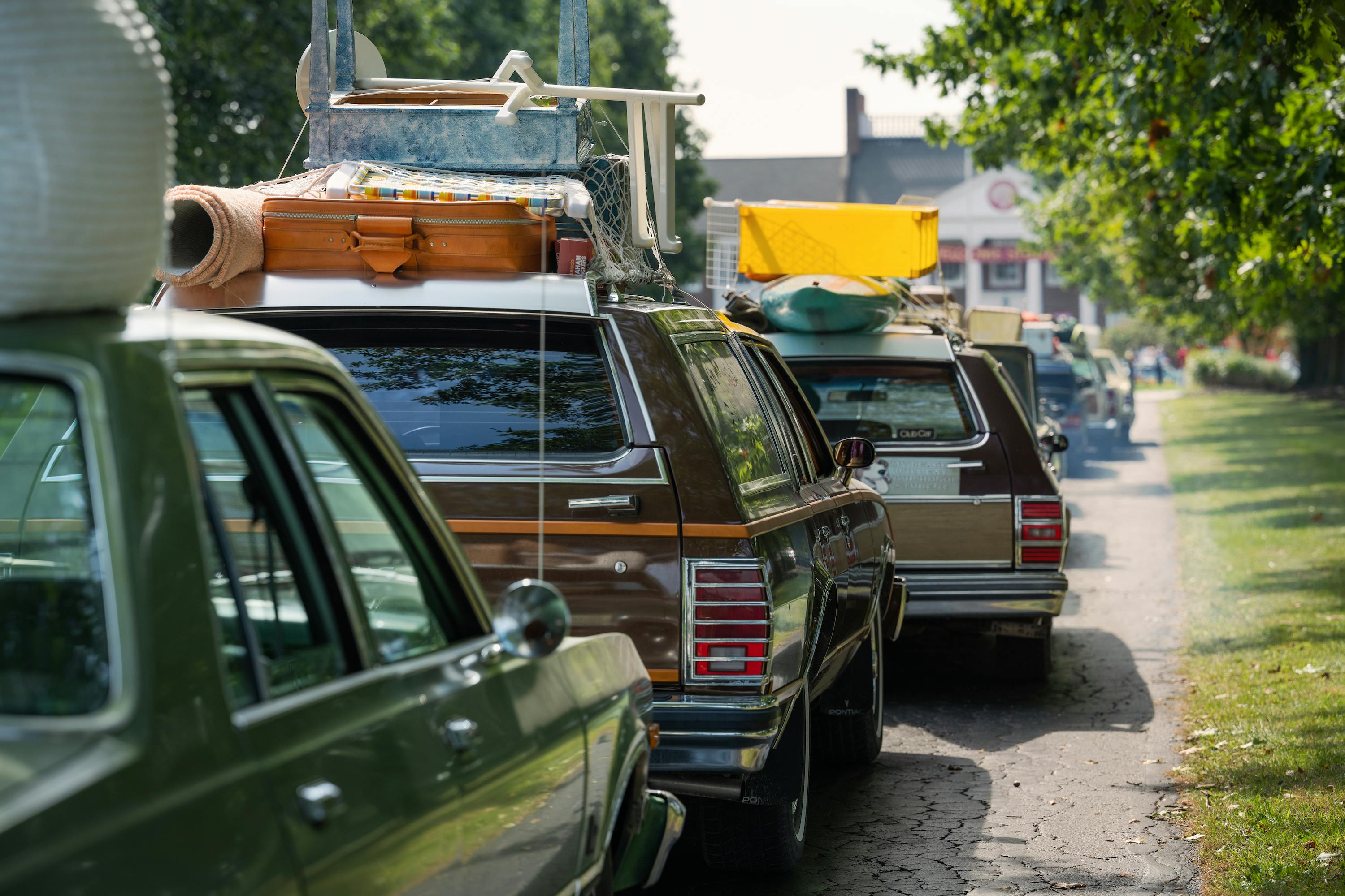 A line of station wagons make their way down a gravel road towards a white college building.