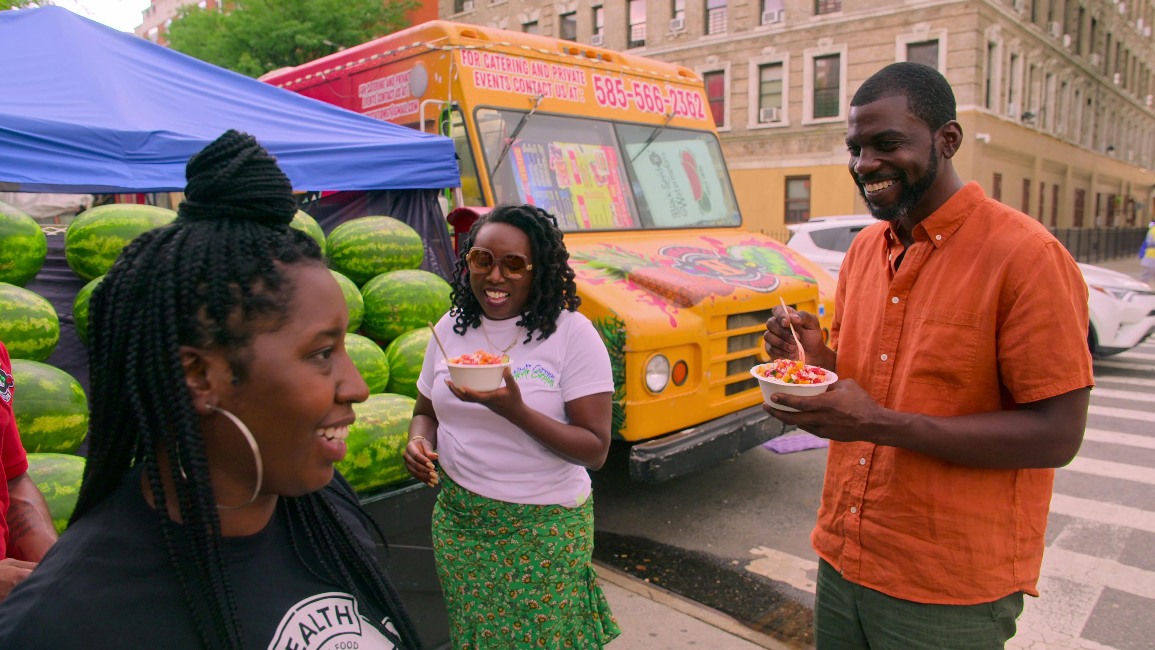Stephen Satterfield eats something from a food truck with two other people. Behind them is a watermelon stand and an orange food truck.