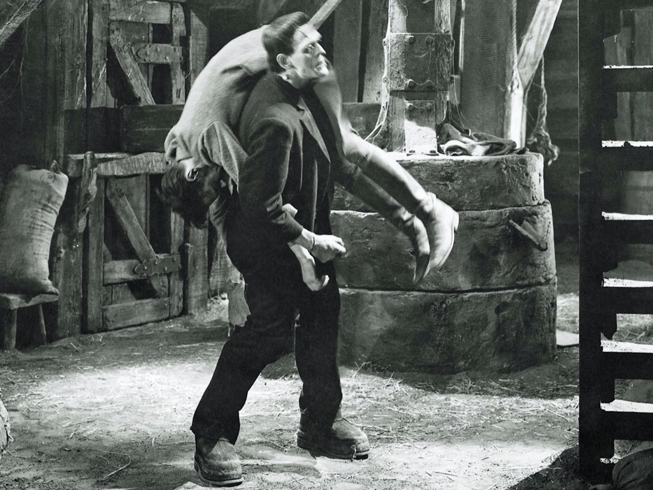 Henry Frankenstein (Colin Clive) in Frankenstein. Frankenstein carries a body over his shoulders through a dusty barn-like building.