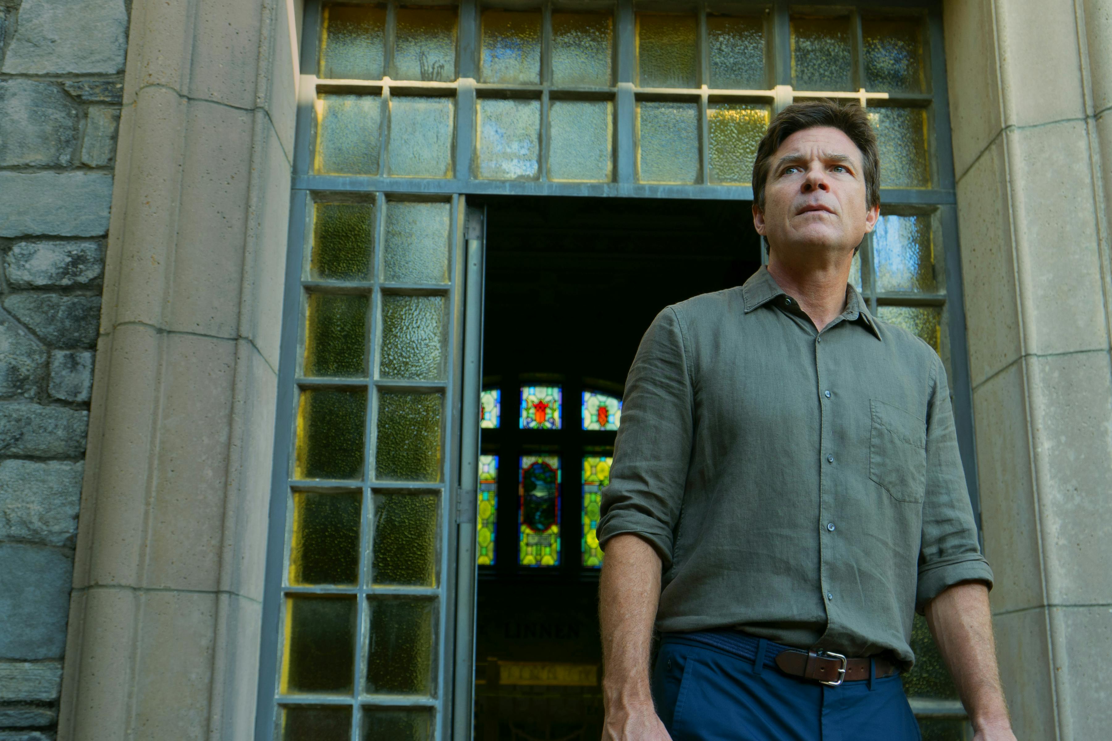Jason Bateman wears a grey buttoned down and jeans and stands outside a glass paned doorway.