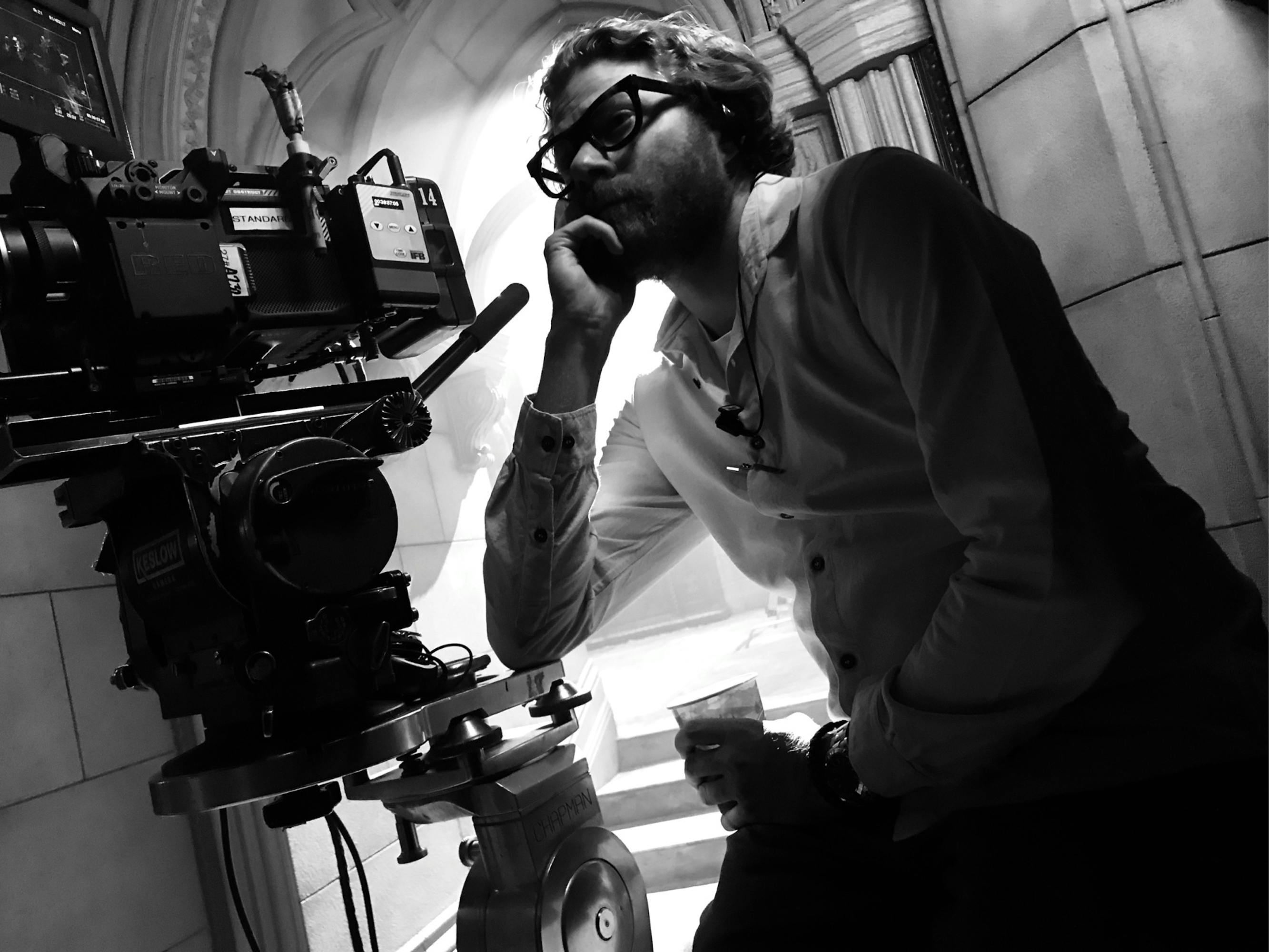 In this black-and-white behind-the-scenes photo, Messerschmidt sits behind a camera, leaning his elbow casually on the stand. He wears square-framed glasses and a white button-down shirt. He’s framed against a stone archway, through which white lights spill from outside.