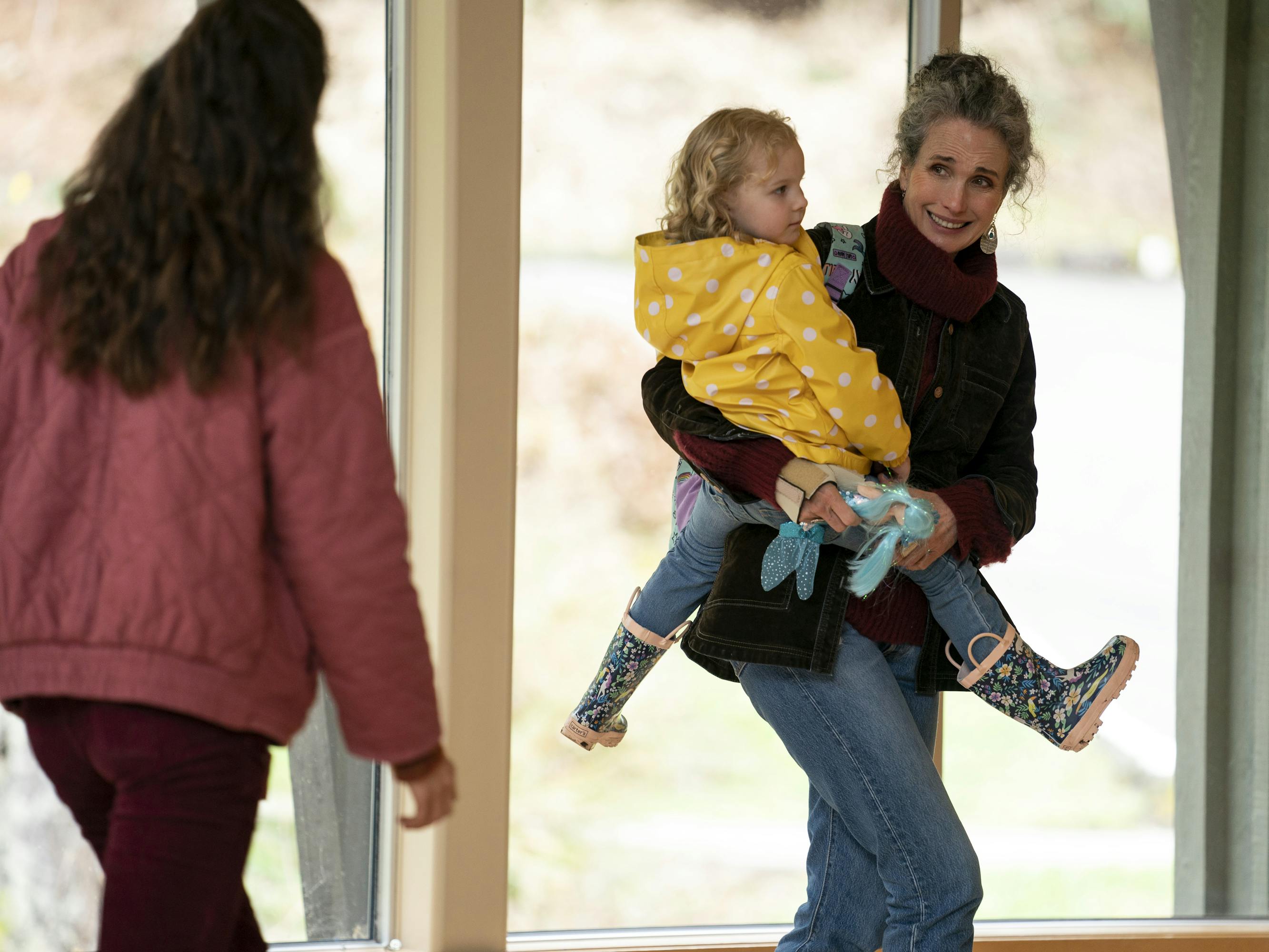 Alex Russell (Margaret Qualley), Maddy Boyd (Rylea Nevaeh Whittet), and Paula Langley (Andie MacDowell) walk through a sun-lit room. Alex wears a pink puffer and red pants, Maddy wears a yellow raincoat and rainboots, and Paula wears a maroon turtleneck.