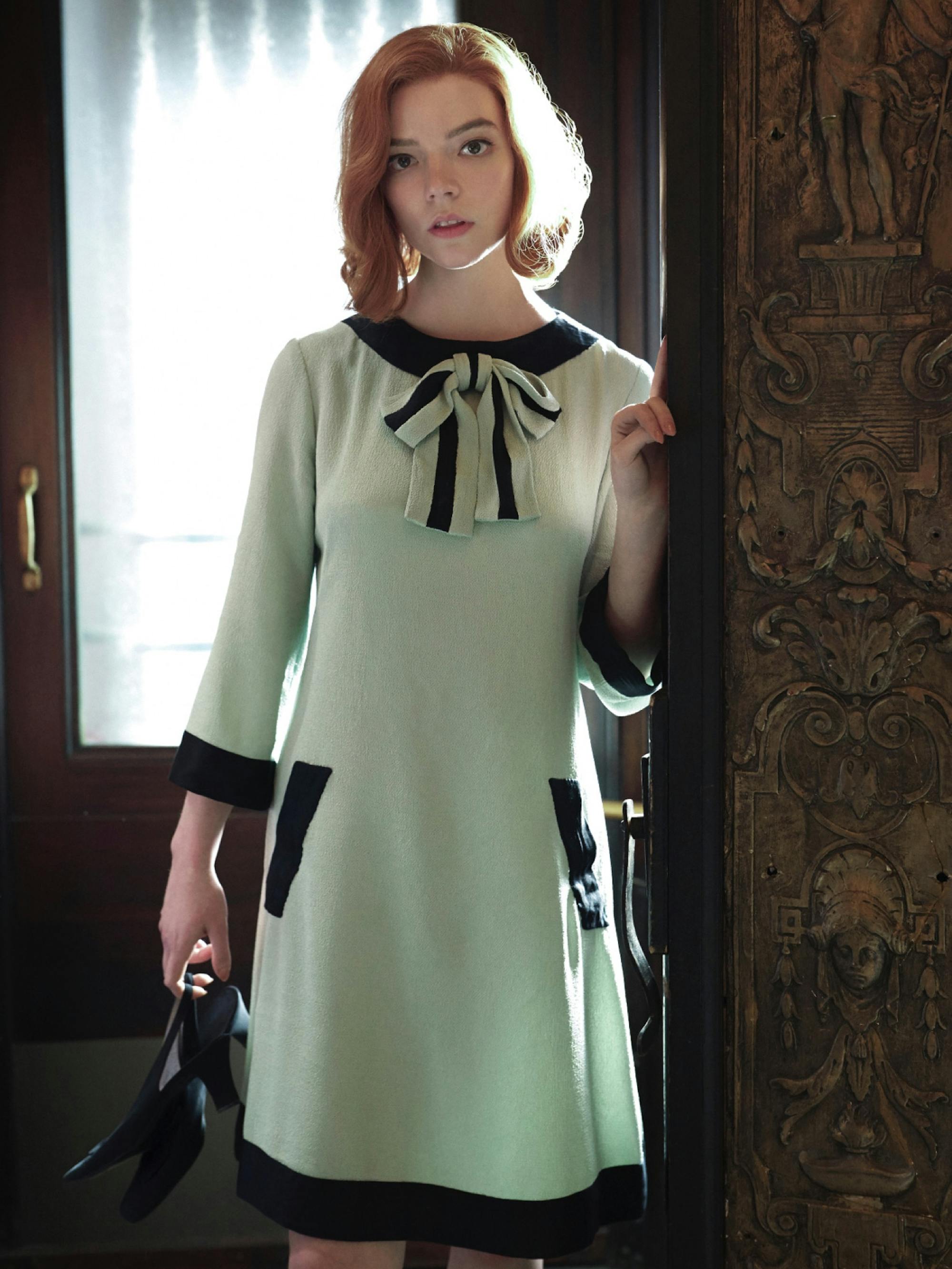 Beth stands in a doorway, heels in hand, wearing her iconic bow dress.