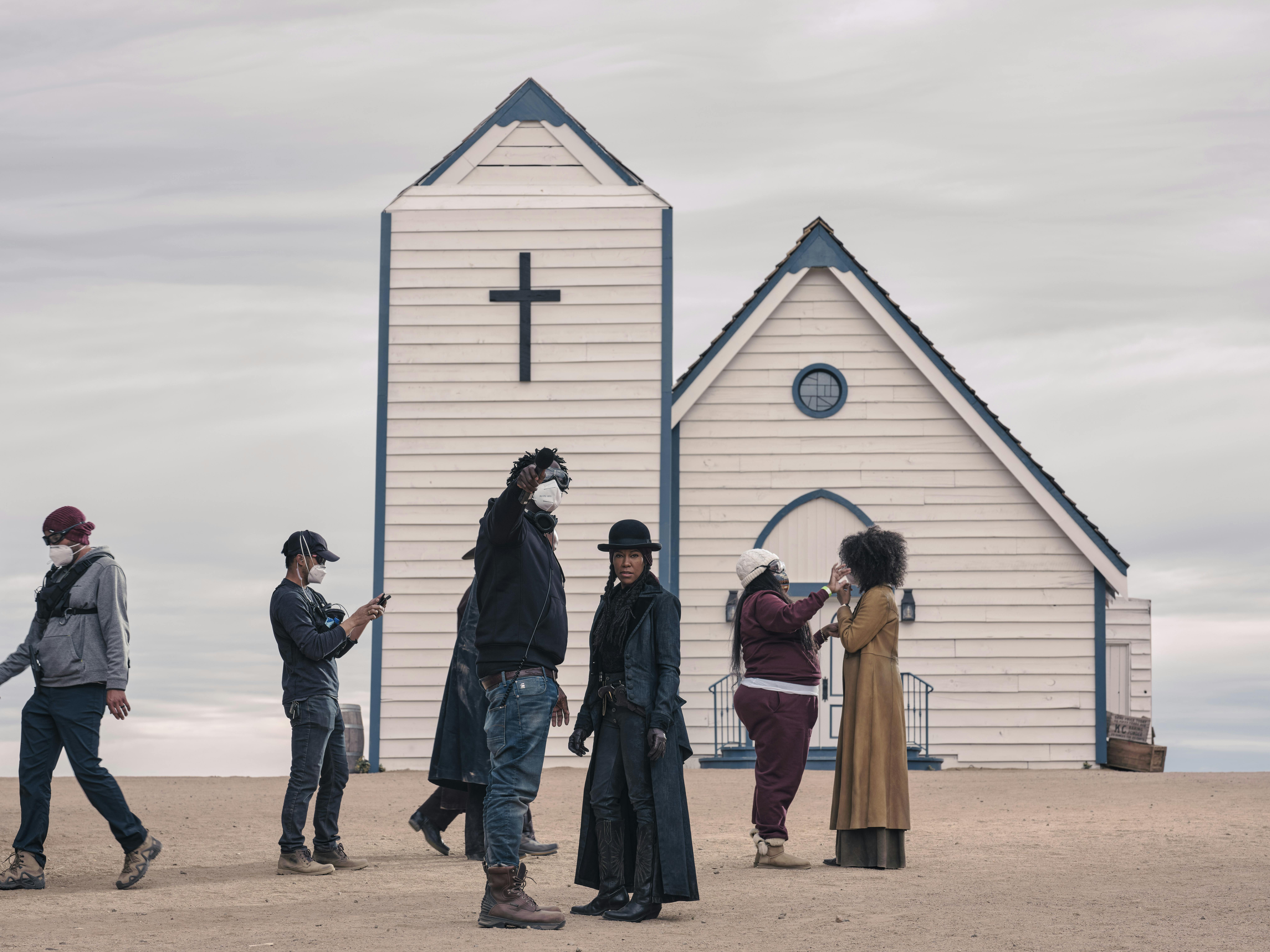 The Harder They Fall cast mingle around the set in costume. Behind them is a white church with a dark cross.