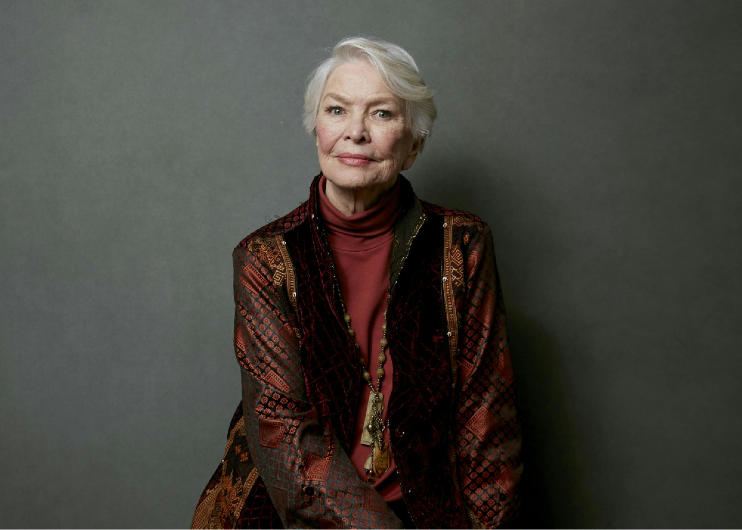 A portrait of Ellen Burstyn, smiling and looking elegant with close-cropped hair and a red jacket.