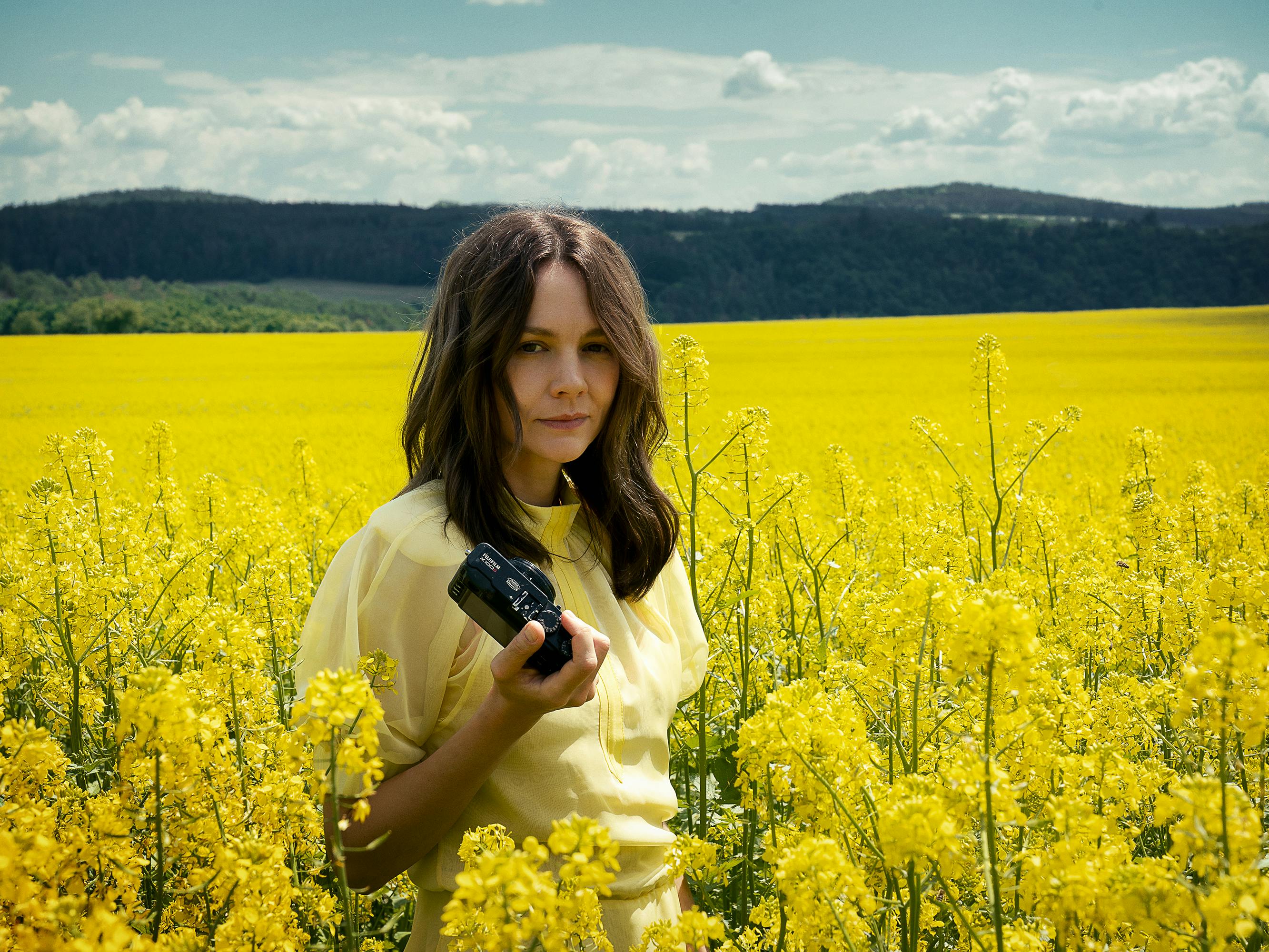 Lenka (Carey Mulligan) wears a yellow top and walks through a field of yellow flowers, holding a camera.