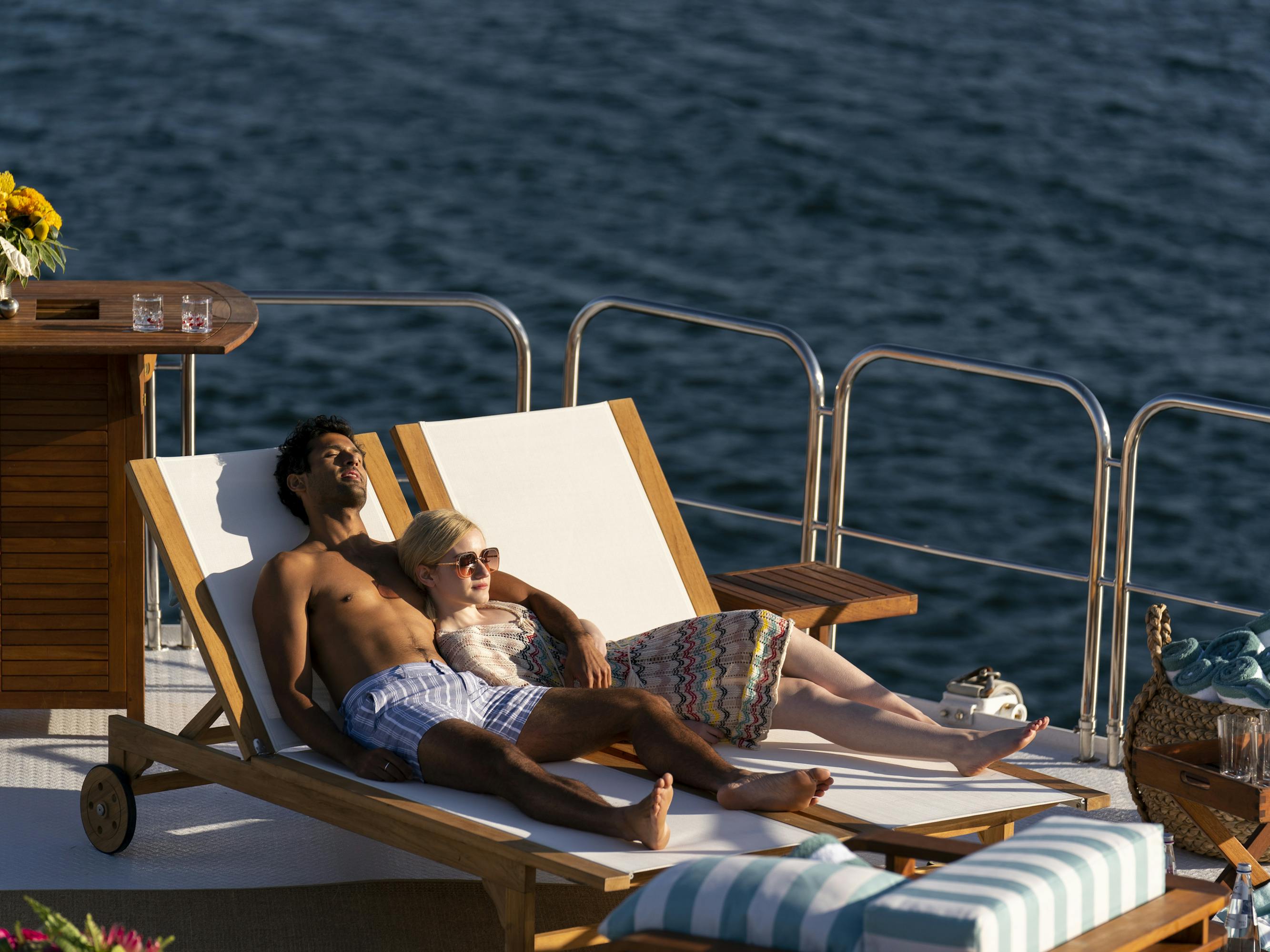 Saamer Usamani and Julia Garner sit on a chaise lounge on a sun-lit yacht. 