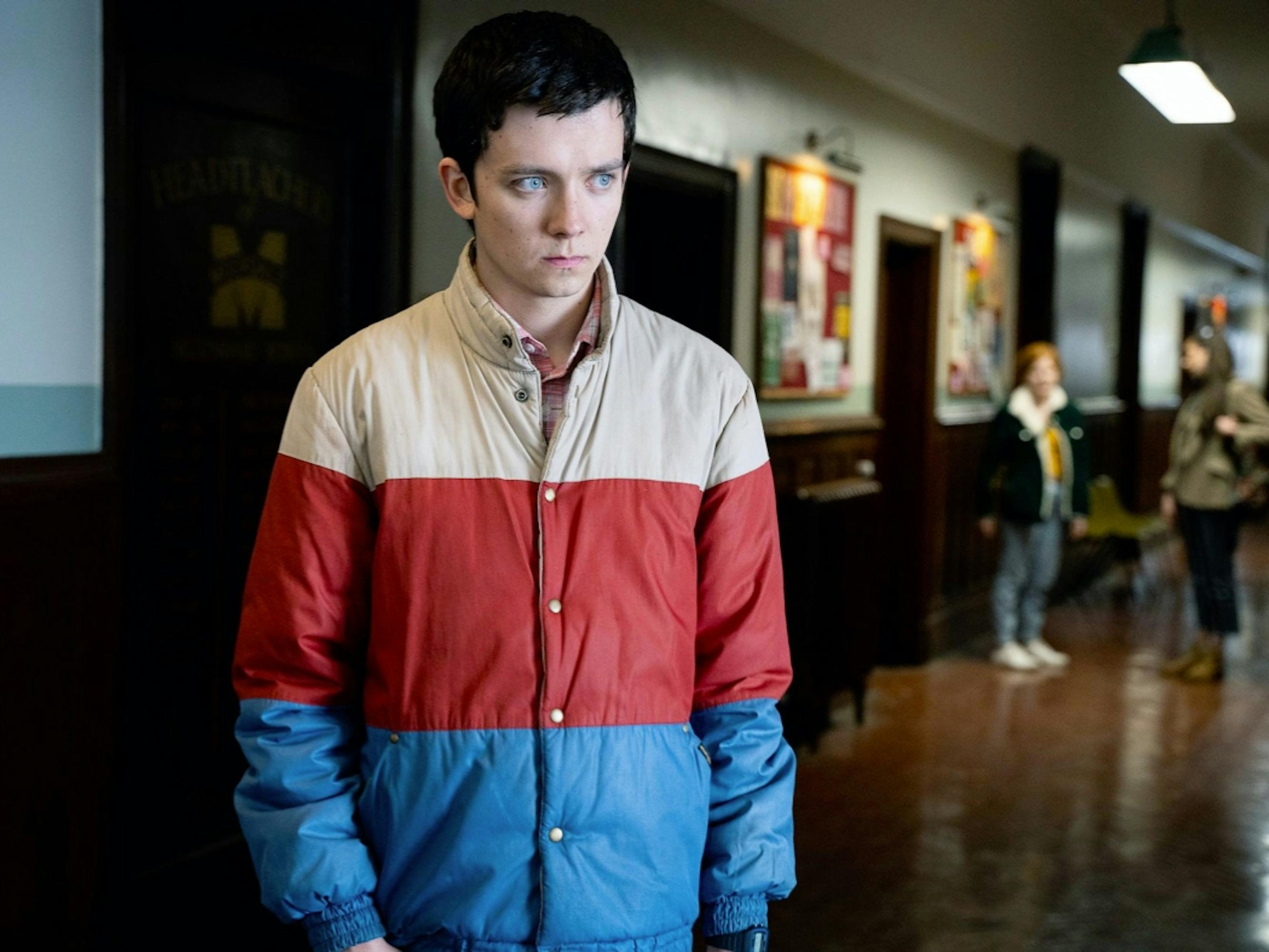 Otis Milburn (Asa Butterfield) wears his tricolored coat as he sulks in the school hallway. The floor is a shiny hardwood, and there are two other students in the distance.
