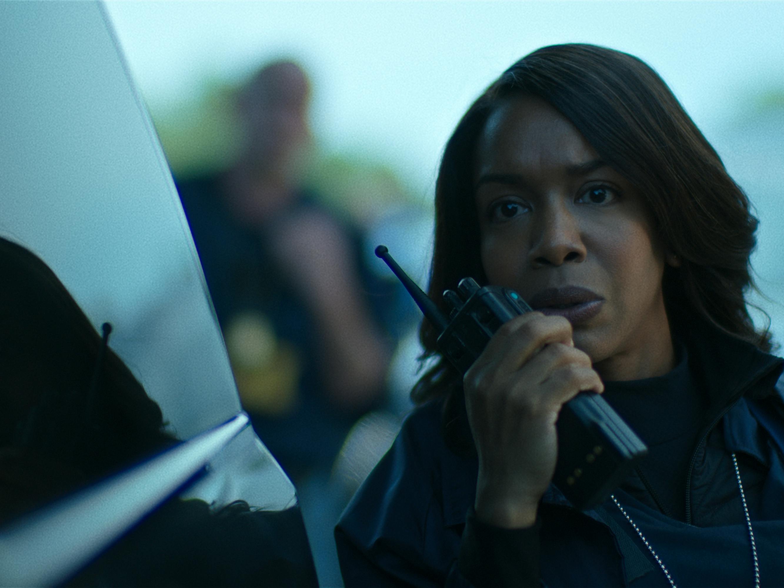 Jessica Frances Duke wears a dark jacket and speaks into a black walkie talkie.