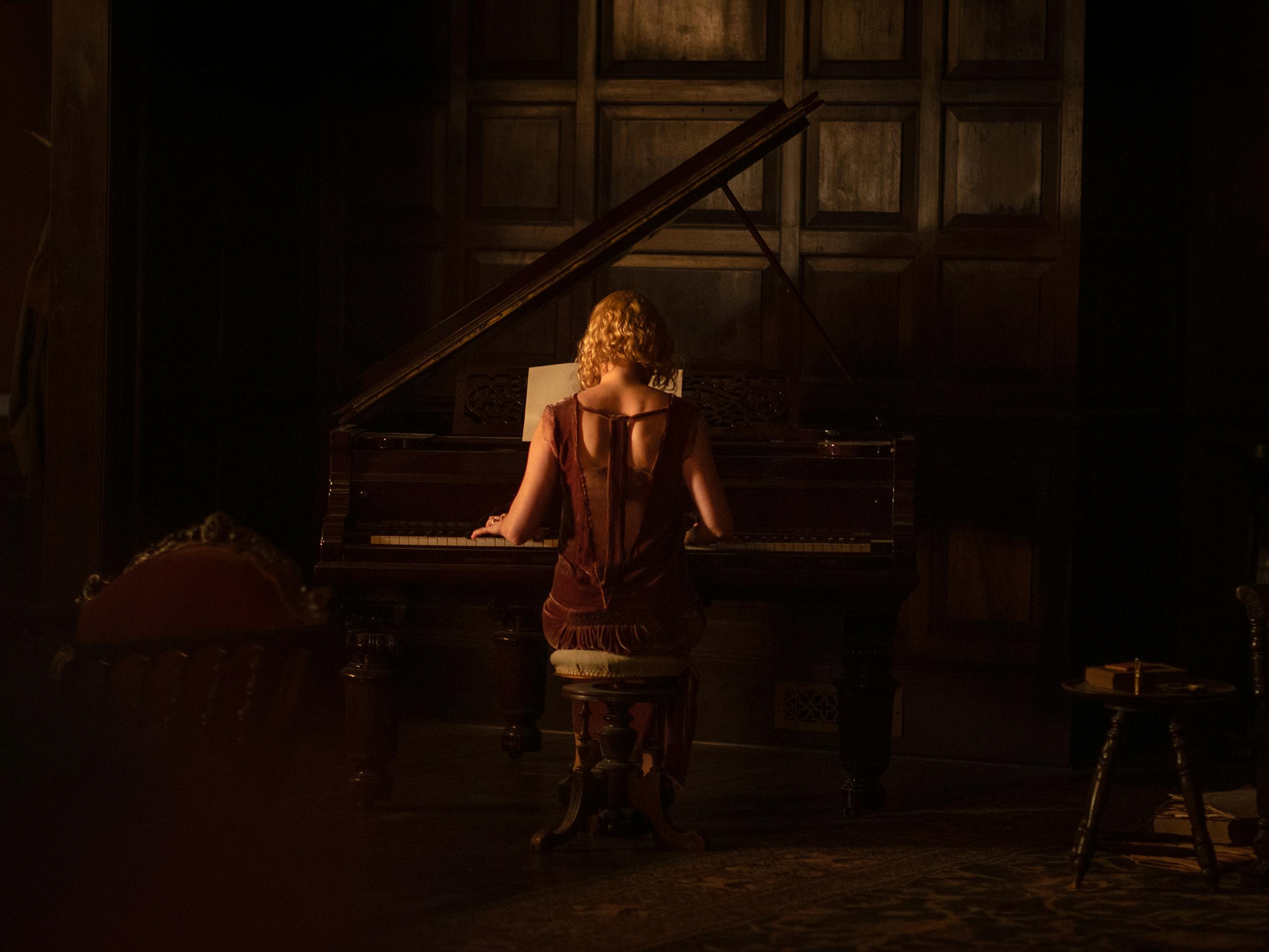 Kirsten Dunst wears a maroon dress and plays piano in a dark room.