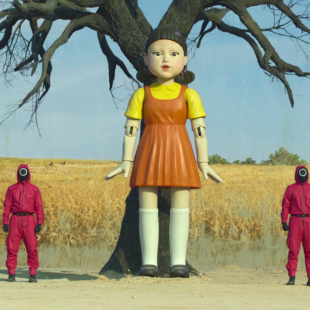 The Squid Game doll is flanked by two red-clothed guards, standing in a field of corn. 