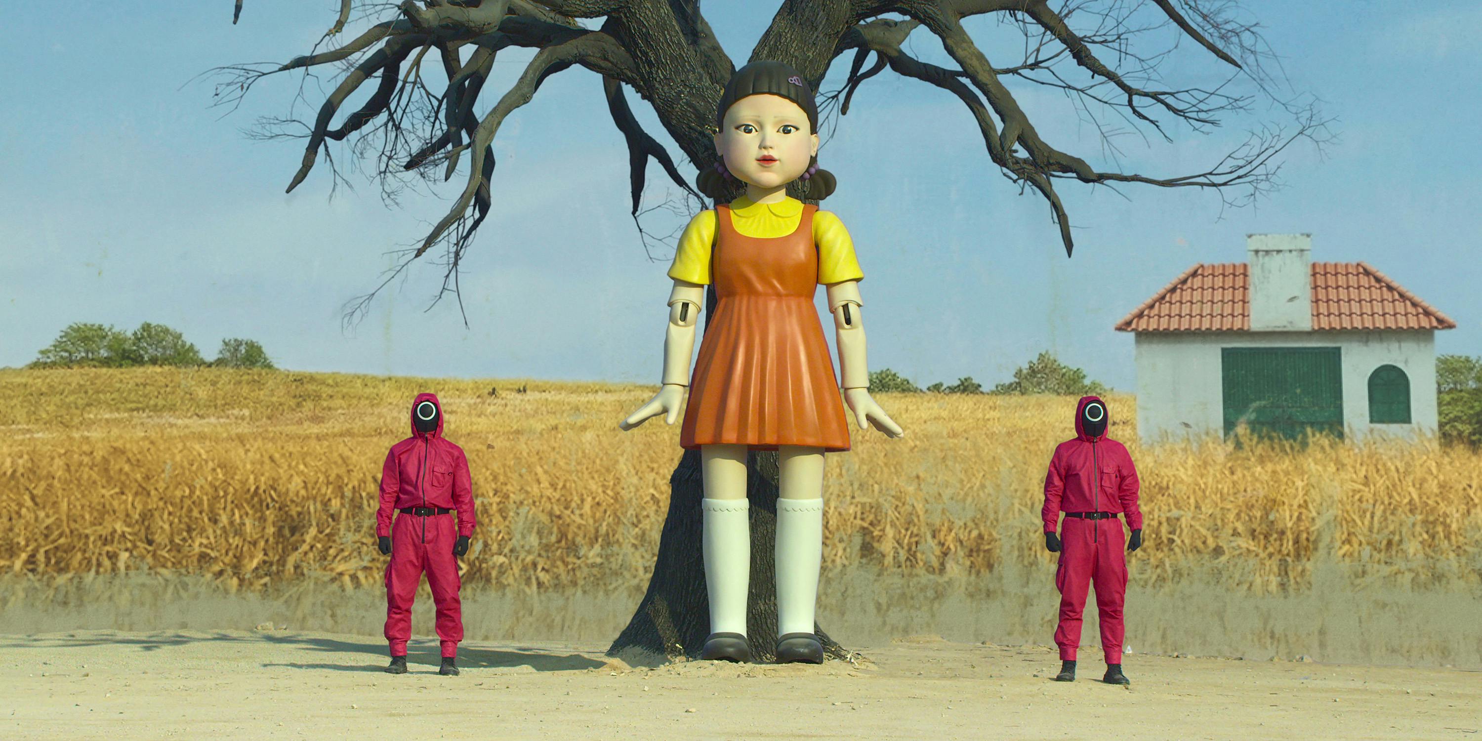 The Squid Game doll is flanked by two red-clothed guards, standing in a field of corn. 