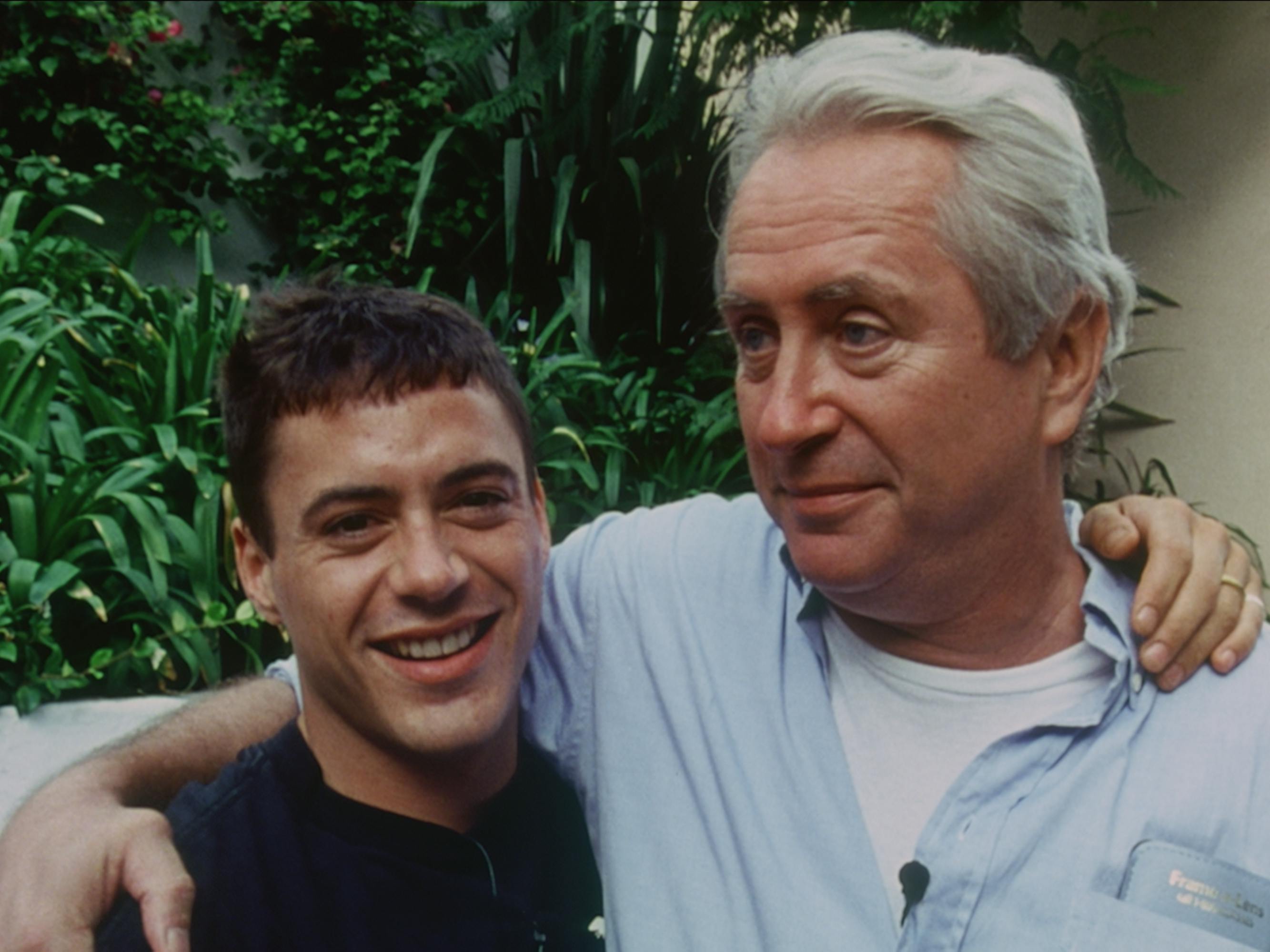A young Robert Downey Jr. stands with his arm around Robert Downey Sr. The former wears a black t-shirt and the latter wears a chambray buttoned-down.