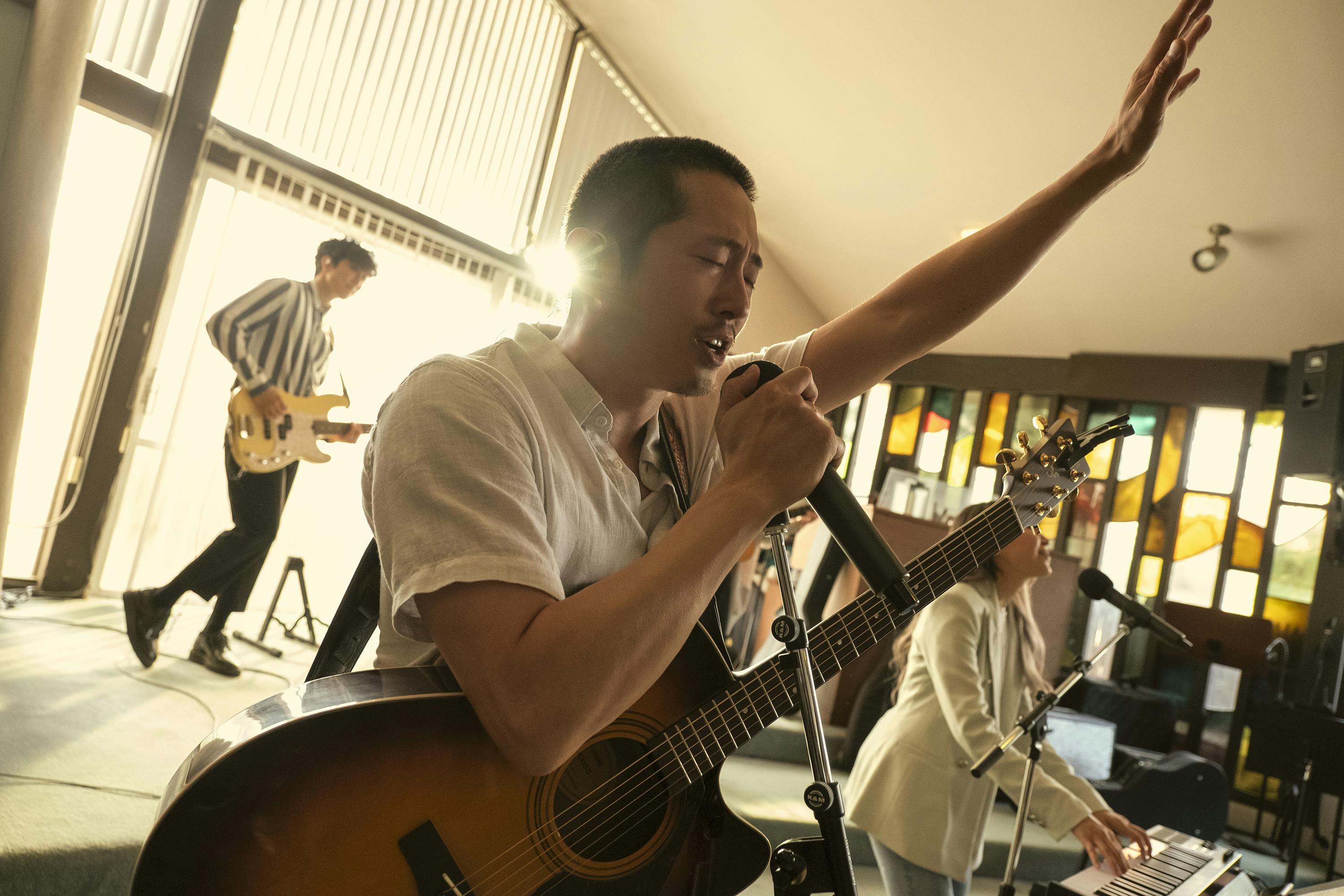 Steven Yeun rocks out in a church. He wears a white shirt and a guitar is slung over his chest. You can almost hear him singing, “That saved a wretch like meeee!”