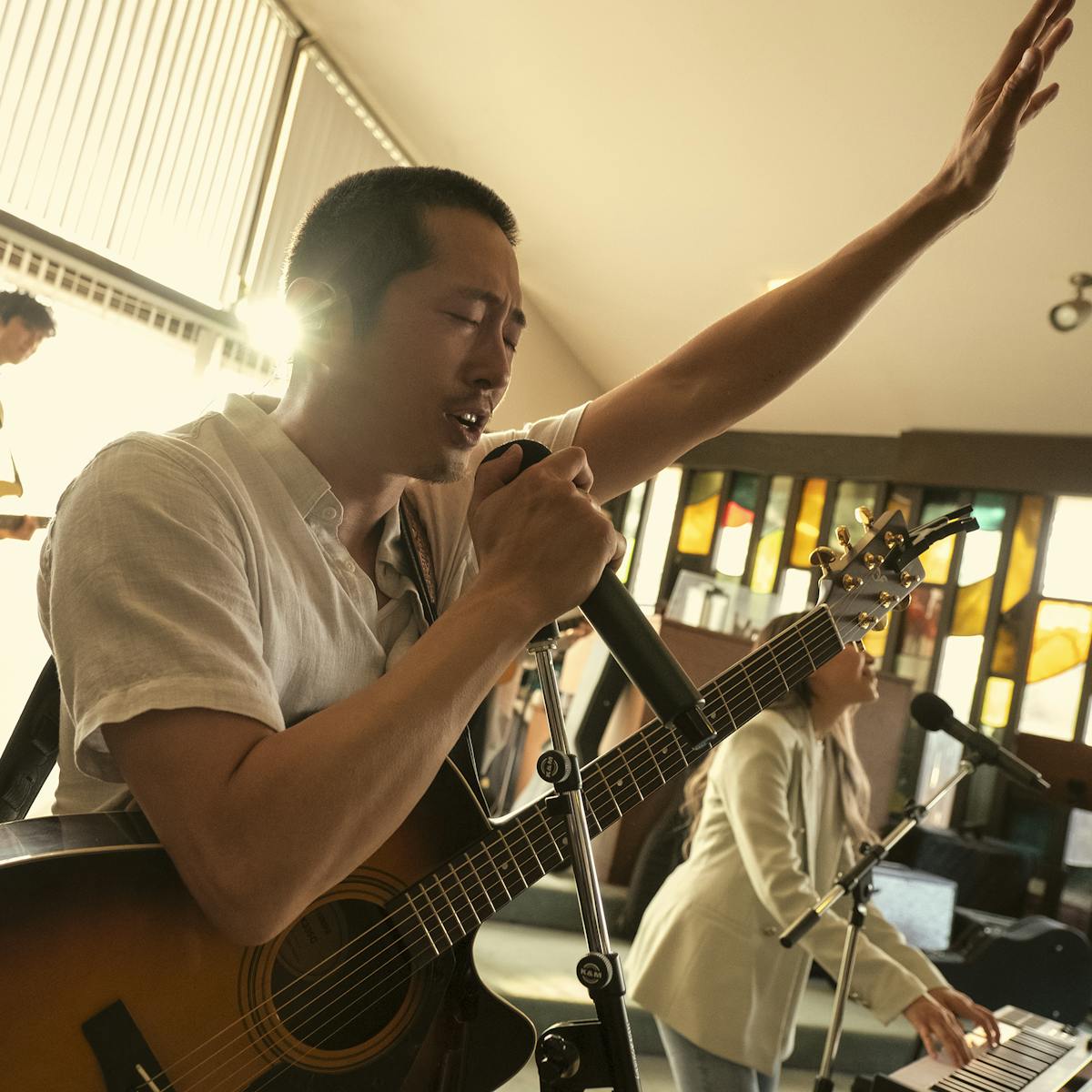 Steven Yeun rocks out in a church. He wears a white shirt and a guitar is slung over his chest. You can almost hear him singing, “That saved a wretch like meeee!”