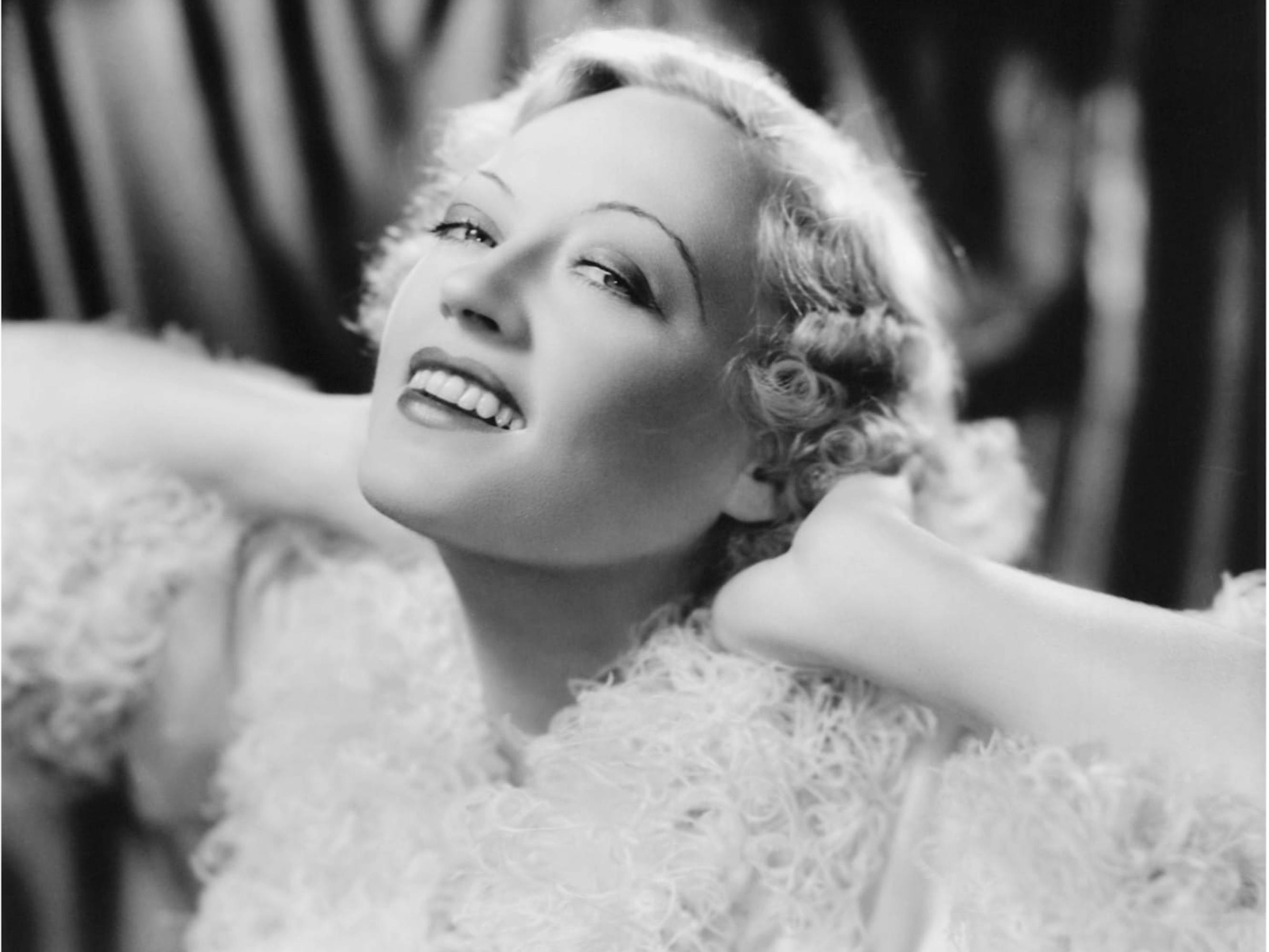 A black-and-white shot of Marion Davies bathed in soft light. She’s framed in front of a curtain, her chin thrown up toward the sky, her arms akimbo as she poses with the back of her head on her hands. She’s wearing a supremely ruffled, light-colored ensemble, and has short, permed blonde hair. A row of straight white teeth is visible through her parted lips and her eyebrows are drawn on in two thin lines.