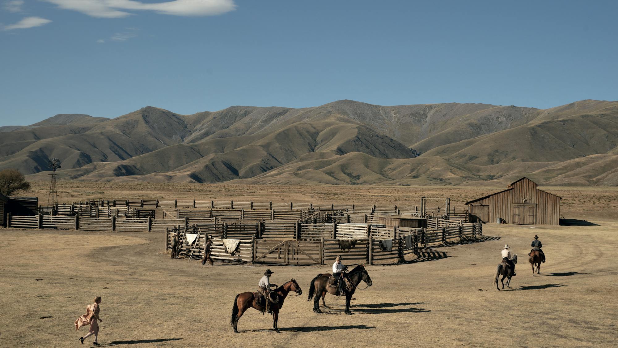 Kirsten Dunst, Kodi Smit-McPhee, Benedict Cumberbatch, and The Power of the Dog cast members ride horses. Kirsten trails behind on her feet. The sky is bright blue, the mountains are shadowed, and there are fences in circular shapes at the enter of the image.