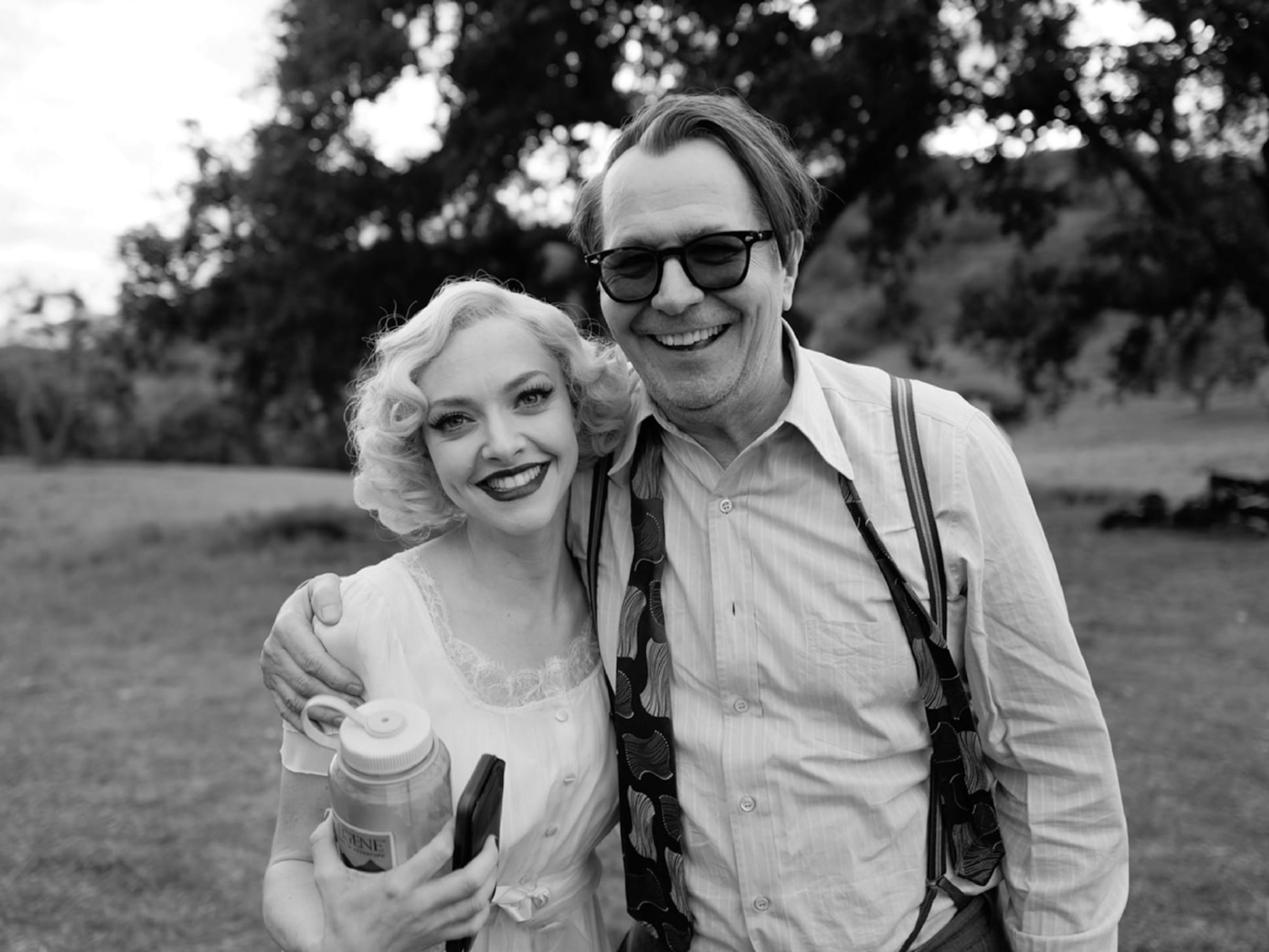 Seyfried and Oldman are pictured in costume outside, with grass, hills, and trees behind them. They have their arms around each other and are smiling. With her blonde hair and lined eyes Seyfried is the picture of Marion Davies, but the cellphone and Nalgene water bottle in her hand give it away.