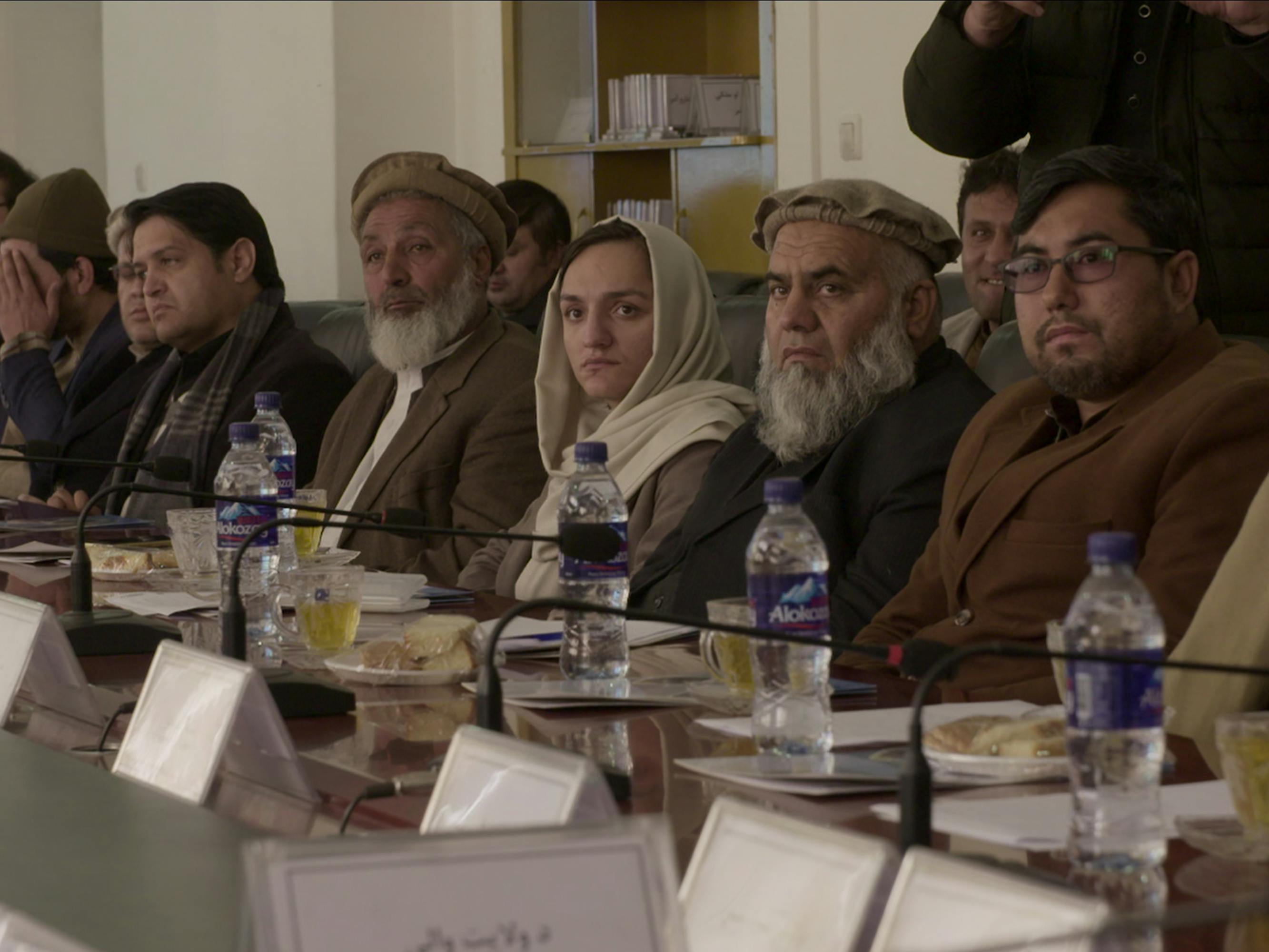 A line of people sit with name cards, water bottles, plates in front of them.