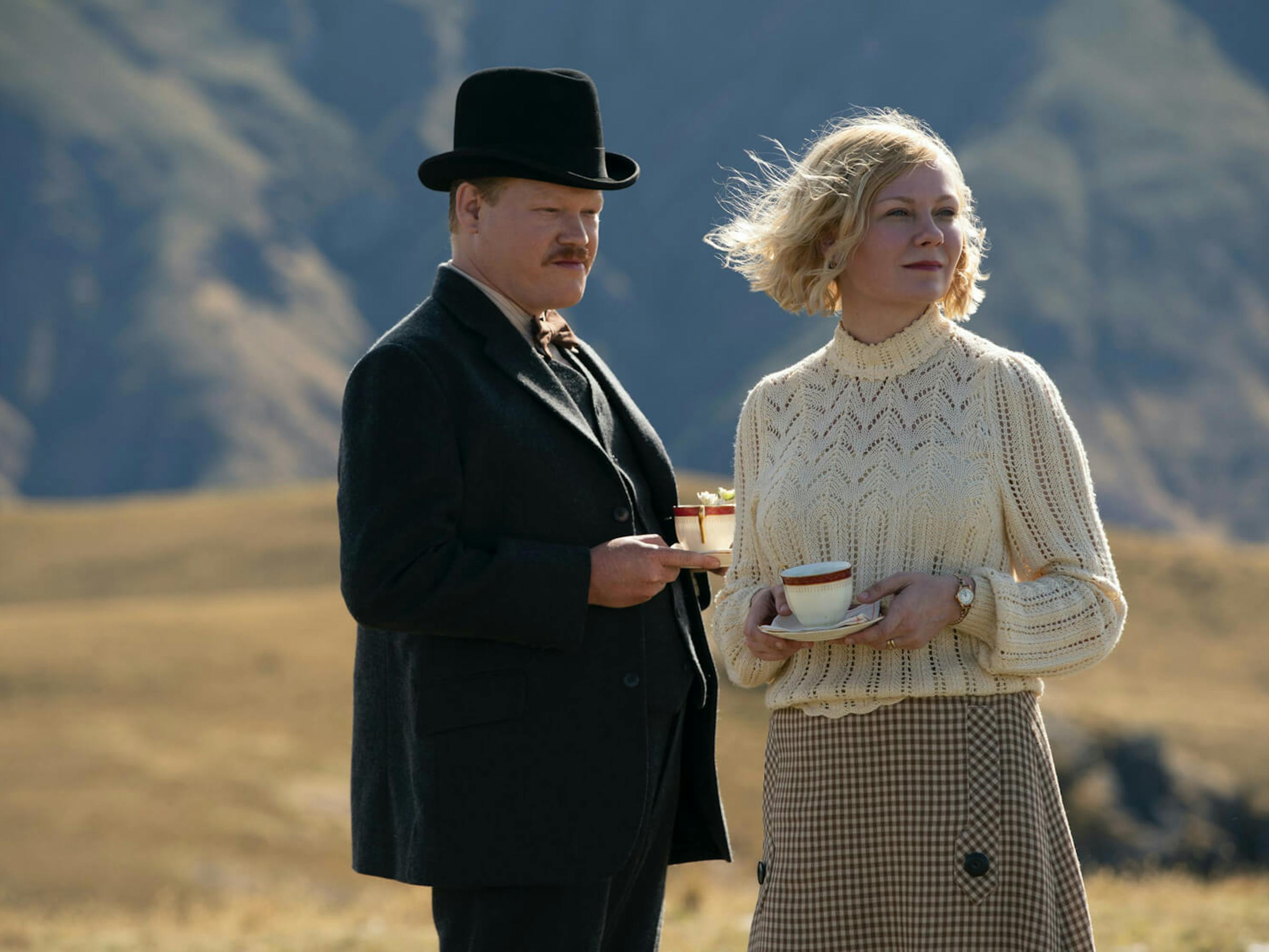 George Burbank (Jesse Plemons) and Rose (Kirsten Dunst) hold teacups and look out on a beautiful windswept scene. Plemons has a mustache and wears a dark suit, bowtie, and a bowler hat. Dunst wears a checkered skirt, and white sweater.