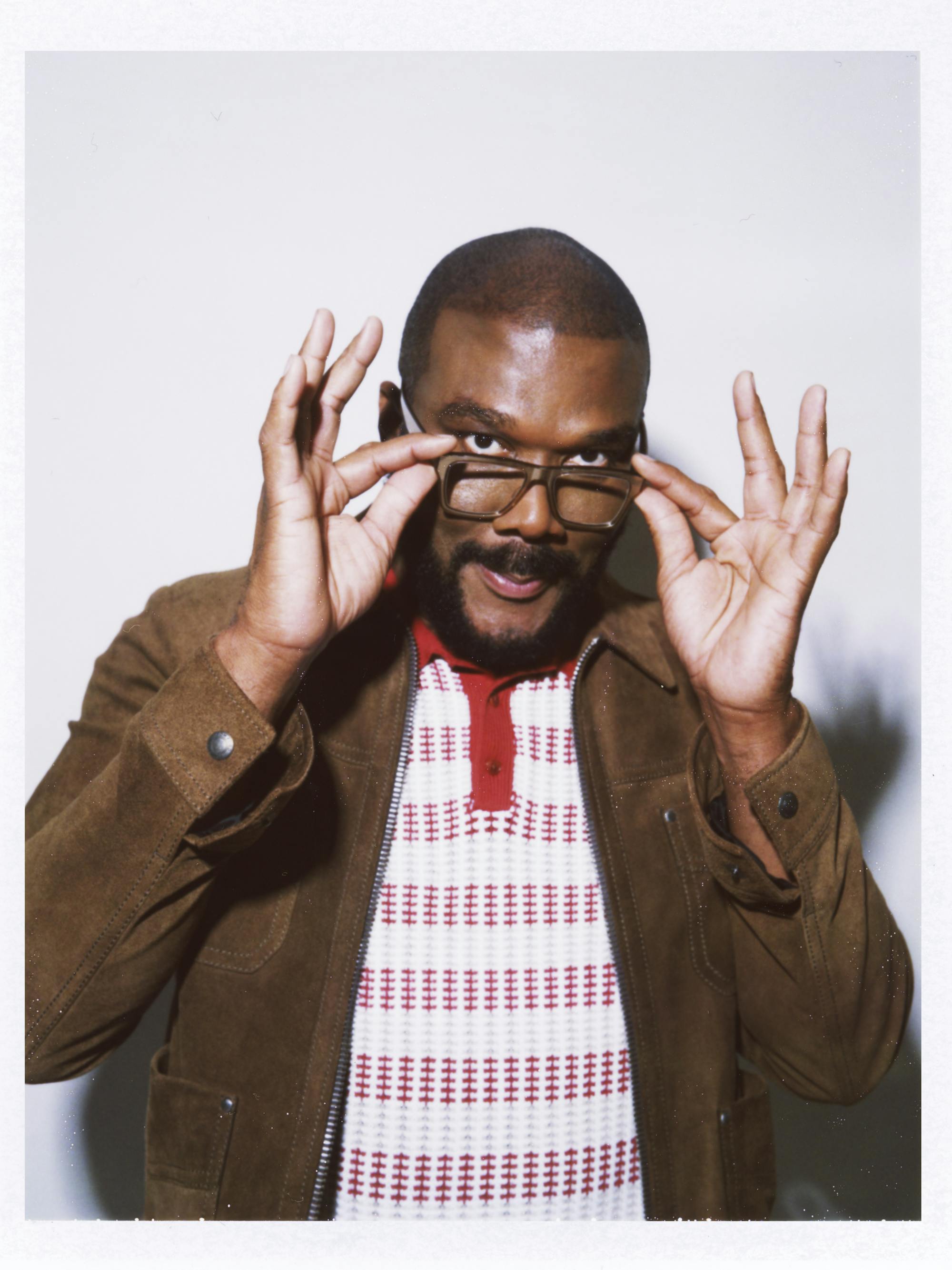 Tyler Perry wears a white and red shirt under a brown suede jacket. He tilts his glasses down at the camera.