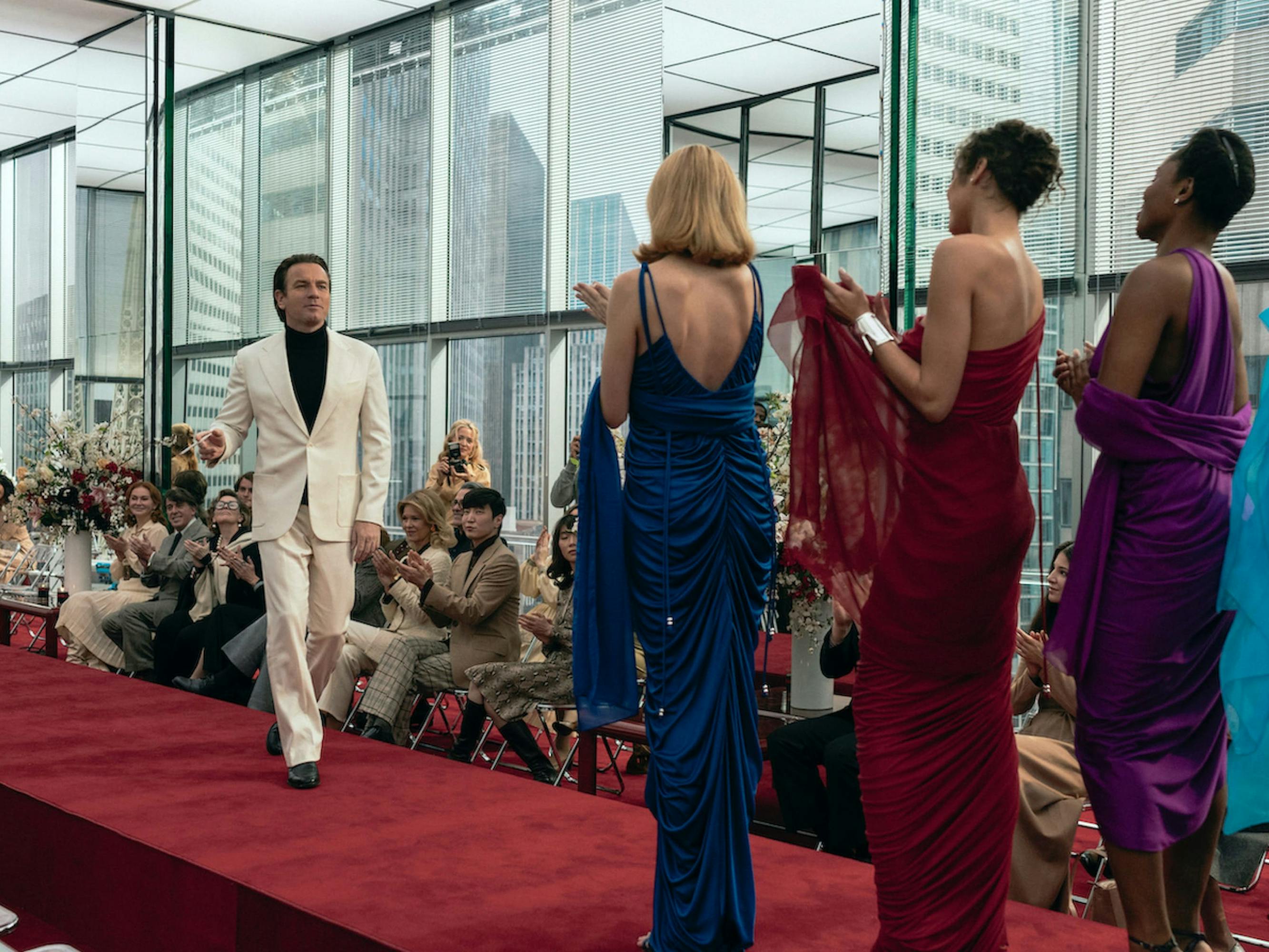 Halston (McGregor) in yet another fierce shot. He struts down a red carpeted runway in an all white suit, with a black turtleneck beneath, smoking a cigarette (he’s consistent!) Everyone alongside the runway claps, and some take pictures. He appears to walk towards four models, in blue, red, purple, and turquoise ruched dresses. The room is bordered by four glass walls that show buildings around them. 