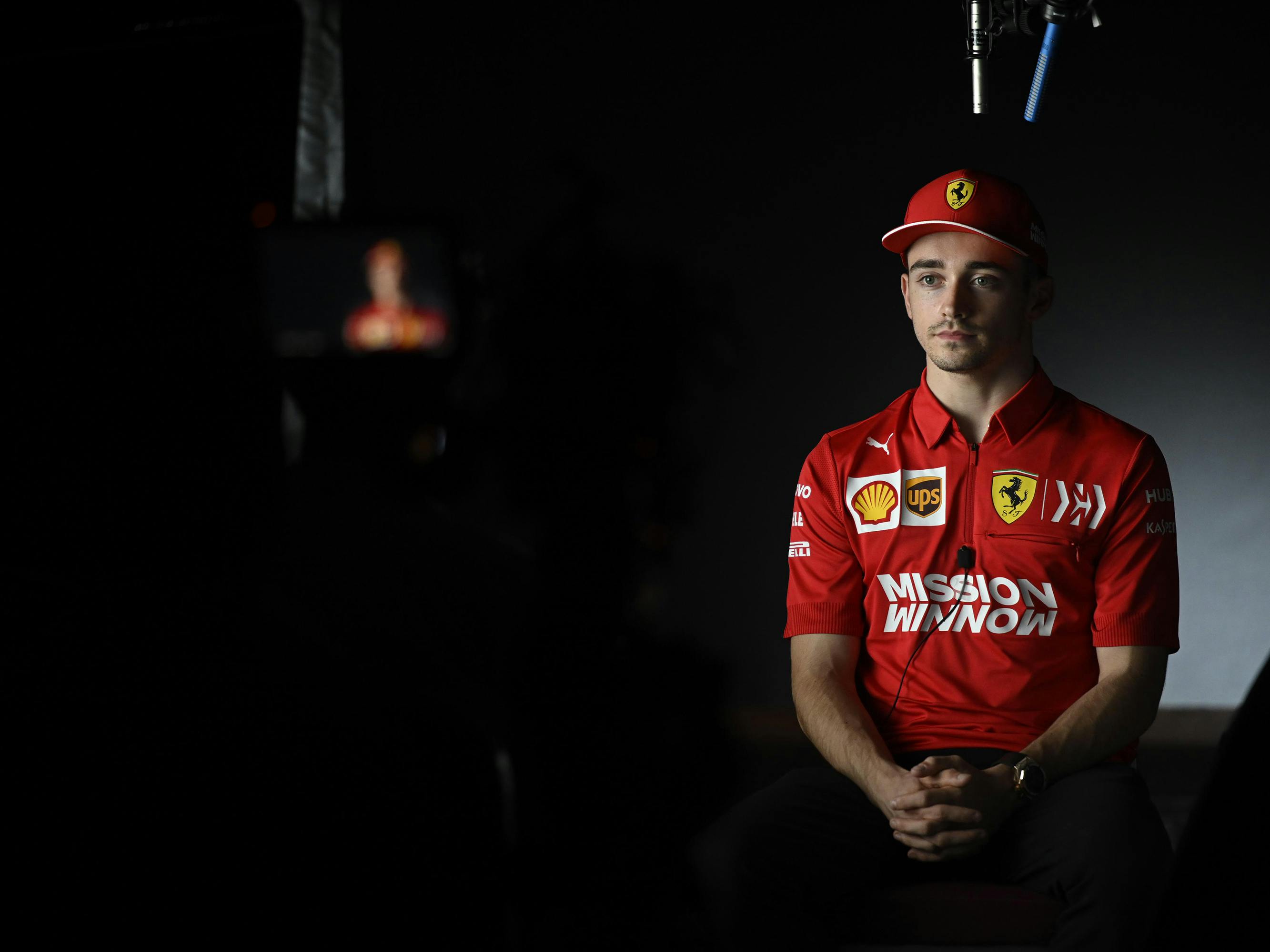 Charles Leclerc wears a red sponsored shirt and hat, and sits politely in the dark.