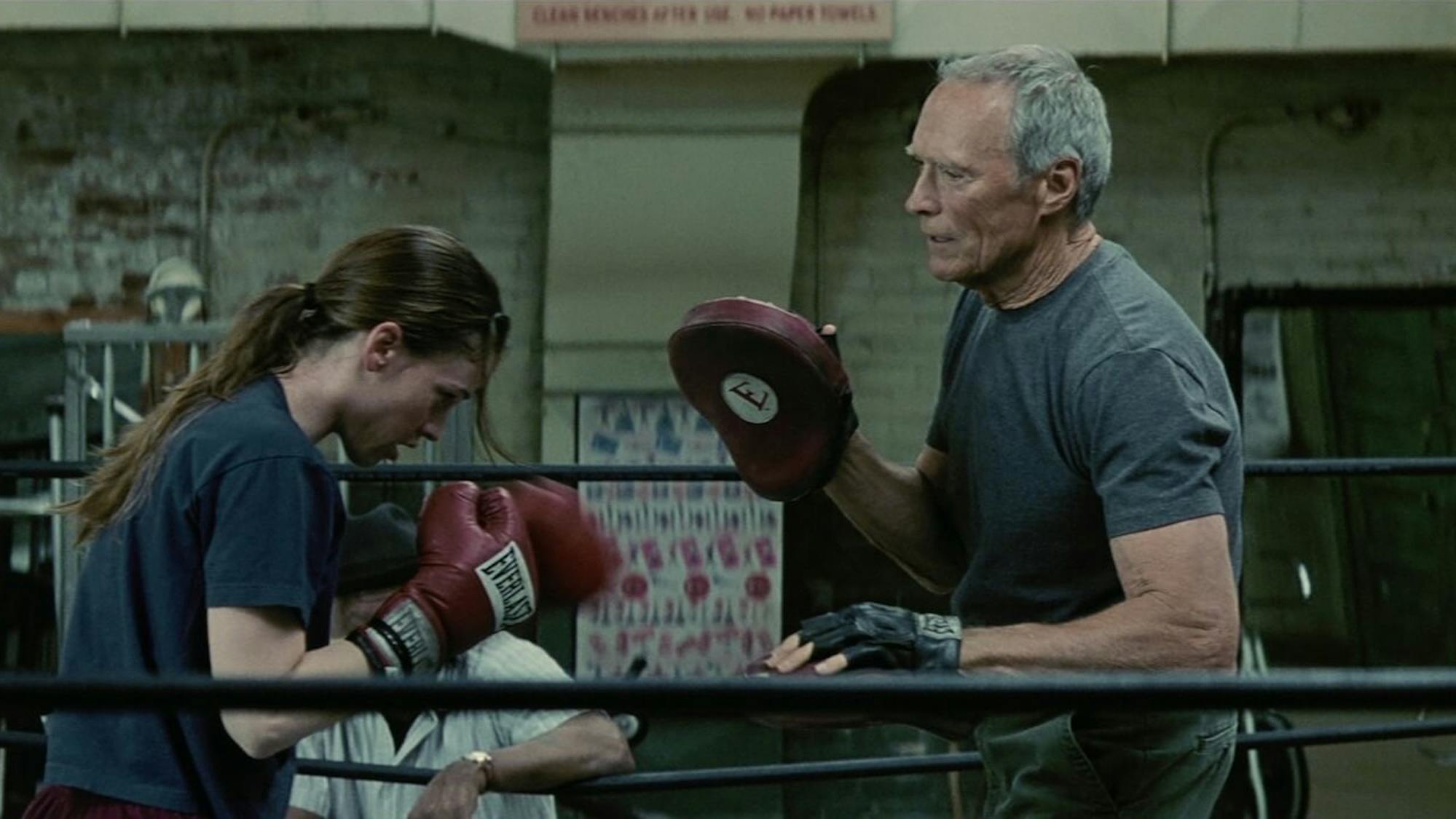 Maggie Fitzgerald (Hilary Swank) and Frankie Dunn (Clint Eastwood) face each other in the gym’s ring. They both wear grey and look exhausted as Maggie gears up to punch Frankie’s glove.
