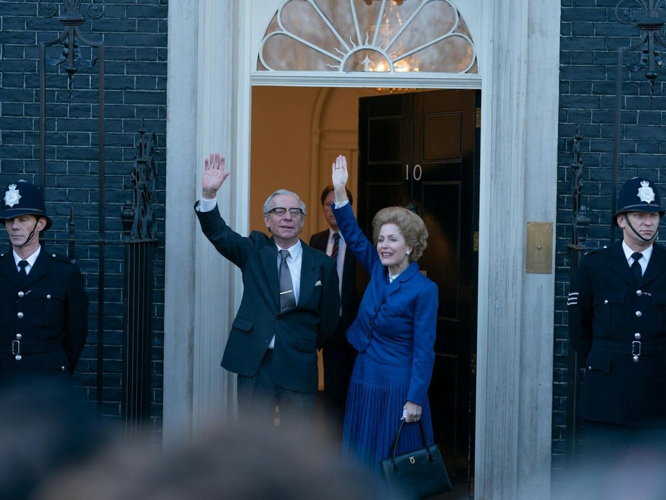 Dennis Thatcher (Stephen Boxer) and Margaret Thatcher (Gillian Anderson) wave to people off camera. Margaret wears a suit in her signature cerulean suit, and their doorway is flanked by two guards.