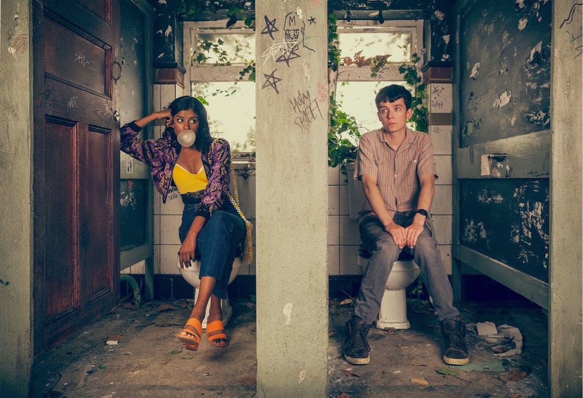 Olivia Hanan (Simone Ashley) and Otis Milburn (Asa Butterfield) in Sex Education. The two characters sit in the abandoned toilets. Ashly wears a chic outfit and blows a massive bubble. Butterfield wears dark jeans and a buttoned down shirt.