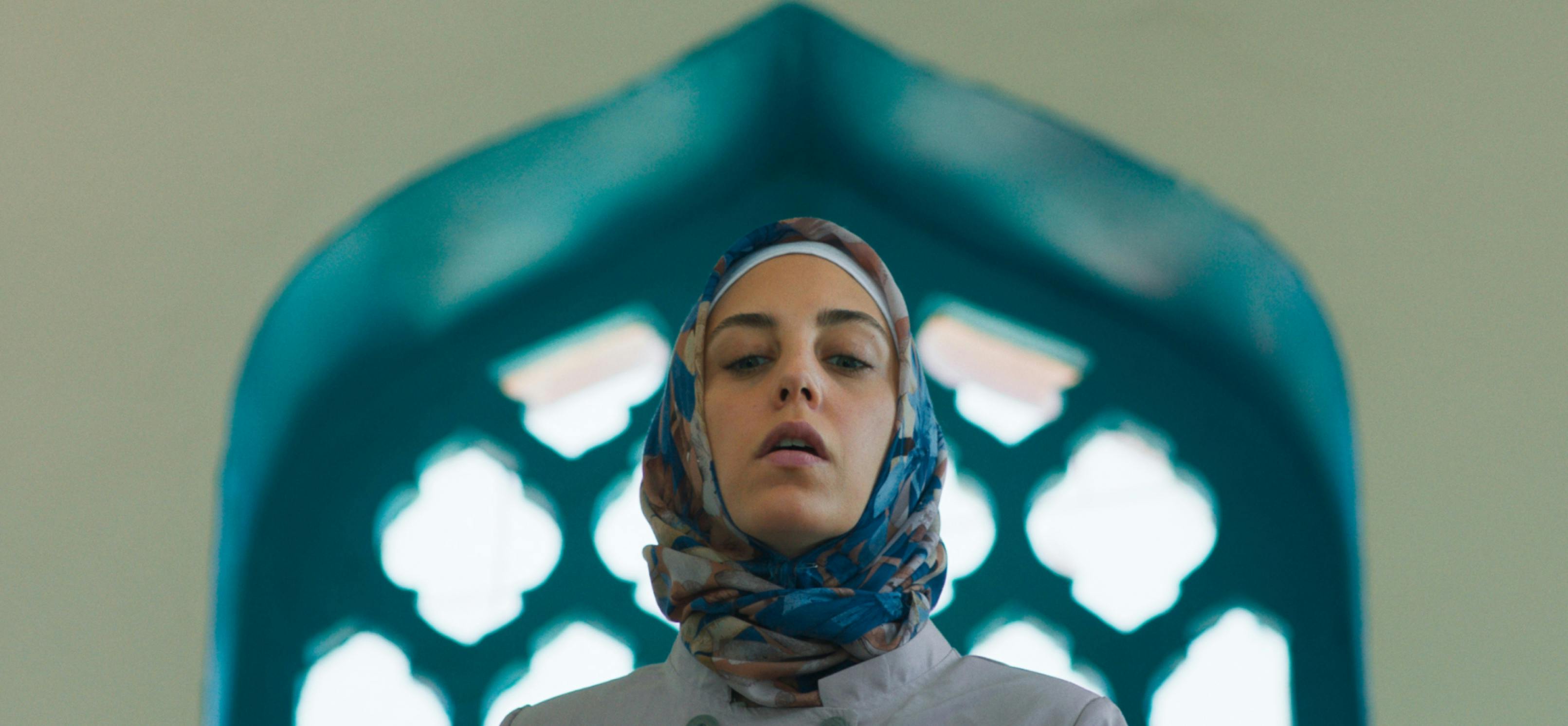 Meryem (actor Öykü Karayel) is framed from the shoulders up against a blue-framed arched window in this shot from Ethos. She wears a headscarf whose turquoise accents blend with the architecture behind her. 