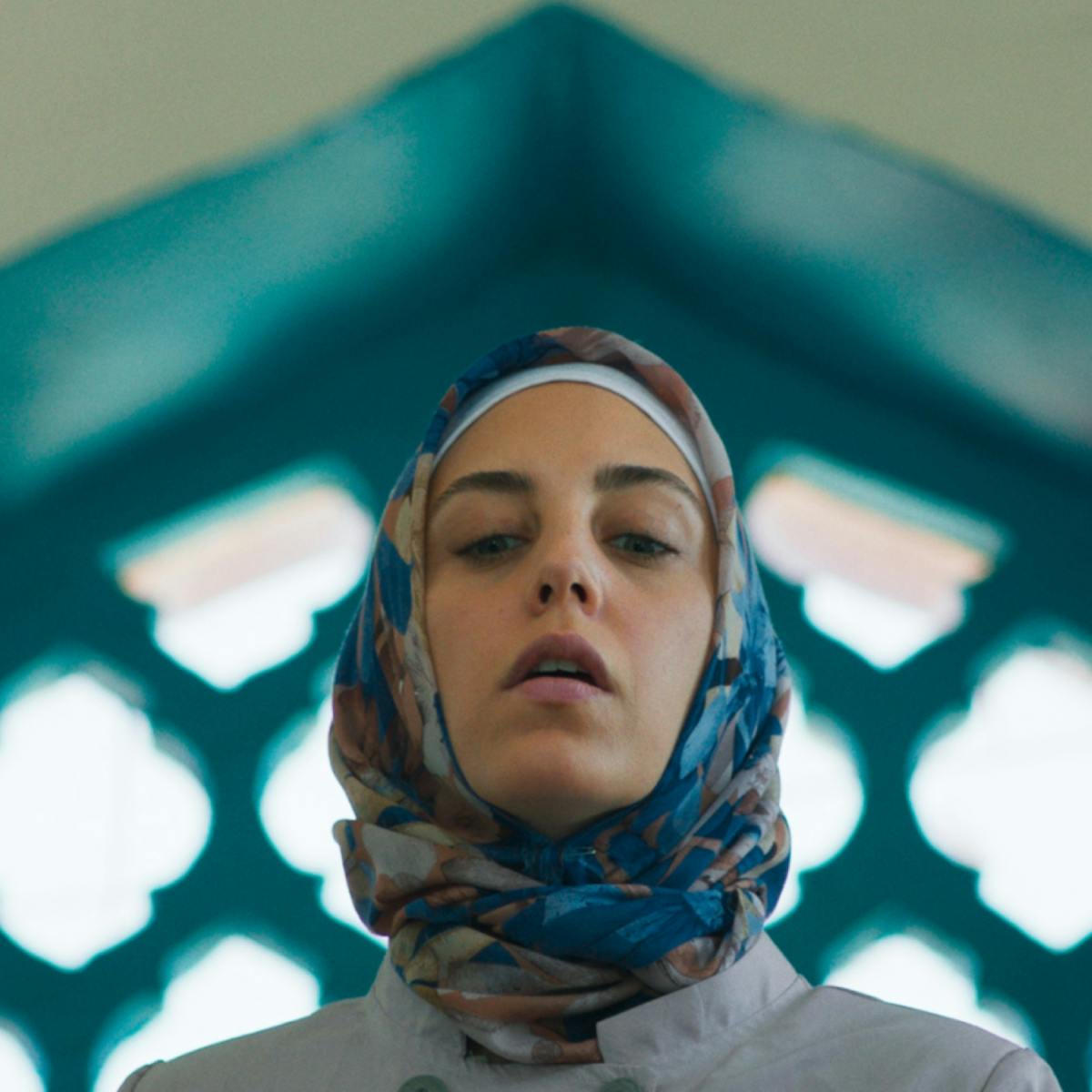 Meryem (actor Öykü Karayel) is framed from the shoulders up against a blue-framed arched window in this shot from Ethos. She wears a headscarf whose turquoise accents blend with the architecture behind her. 