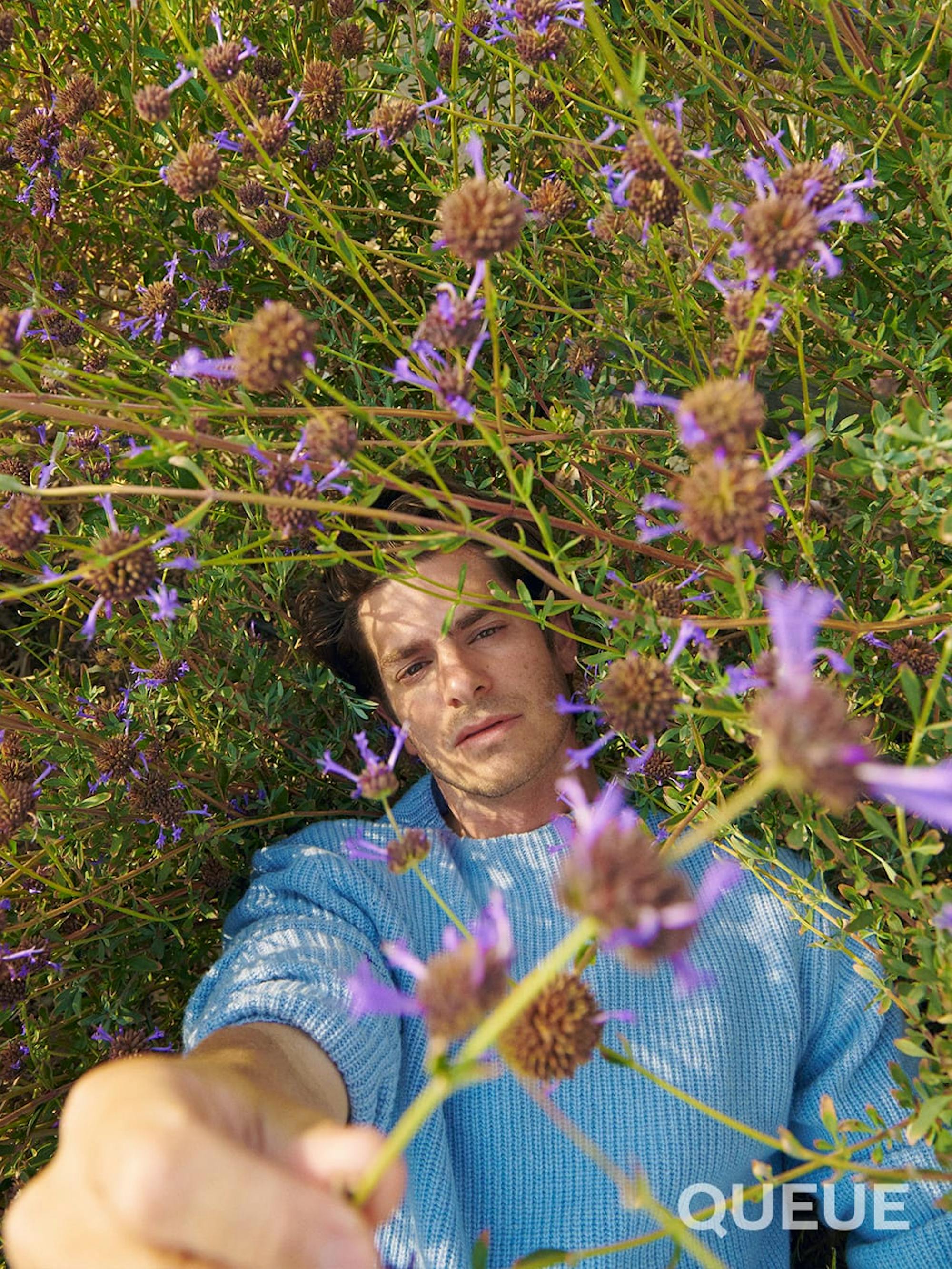 Andrew Garfield wears a blue sweater and lies in a bush of purple flowers. He smizes at the camera.